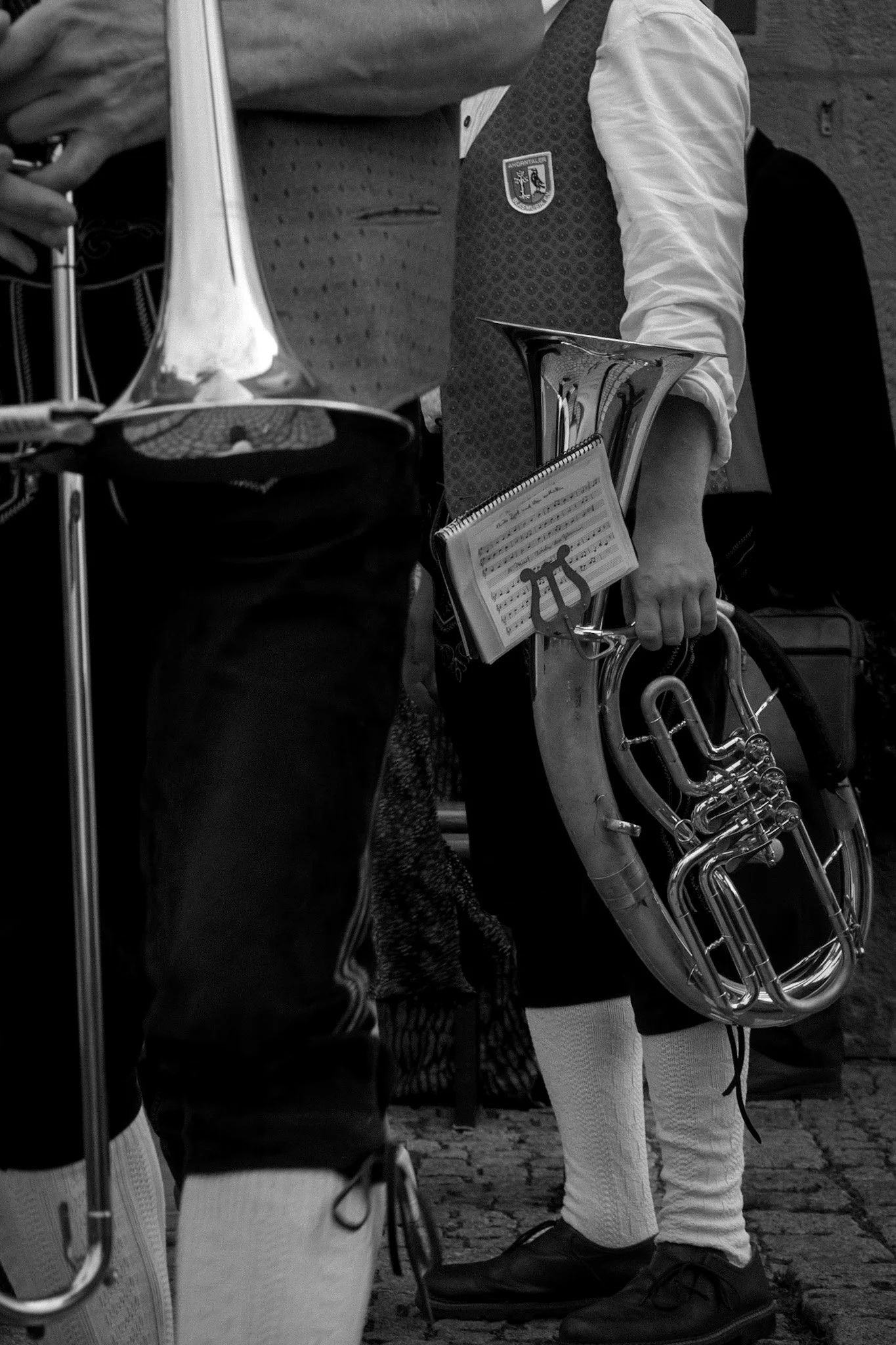 A close-up view of a person holding a trumpet, with another person next to them wearing dark pants and shoes, standing on a cobblestone ground. Both individuals are dressed in formal attire, with one person wearing a jacket with a crest and the other