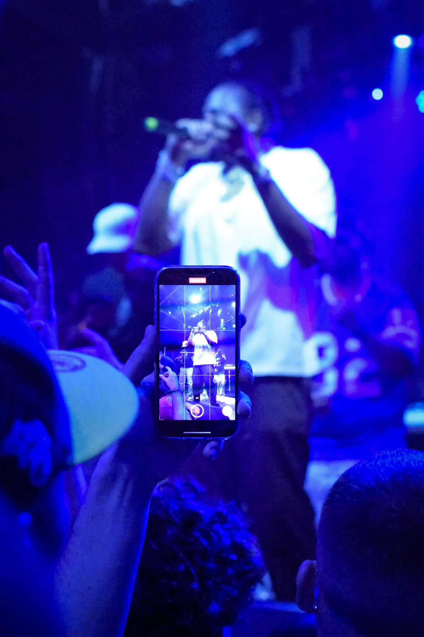 A performer on stage wearing a white shirt and glasses, holding a microphone, with blue and purple stage lighting. Audience members are taking photos and videos with their phones.