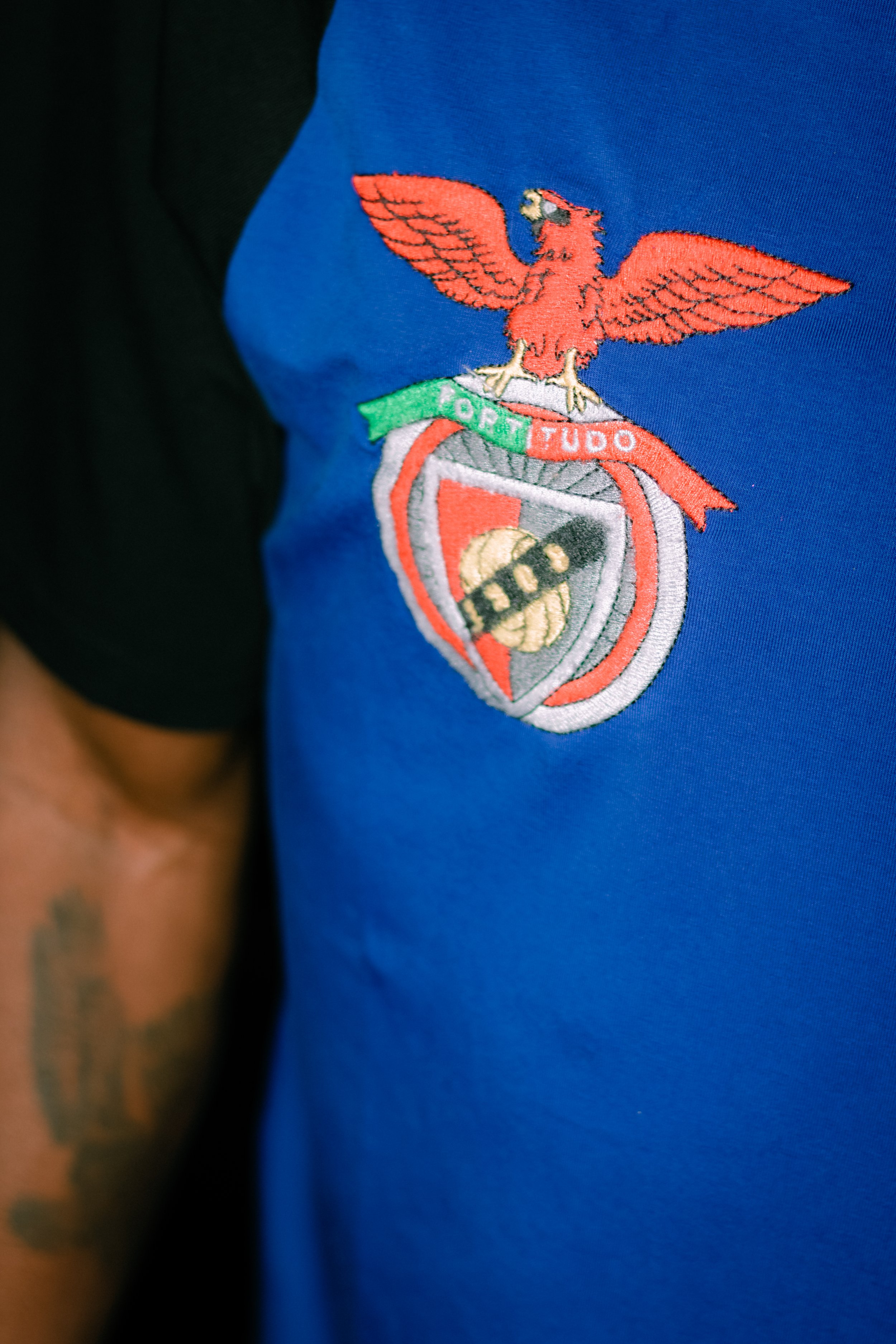 Close-up view of an embroidered emblem on a blue garment. The emblem features an orange eagle with outstretched wings, perched above a shield with a red cross, a golden football, and black elements. A green and red banner beneath the eagle displays t