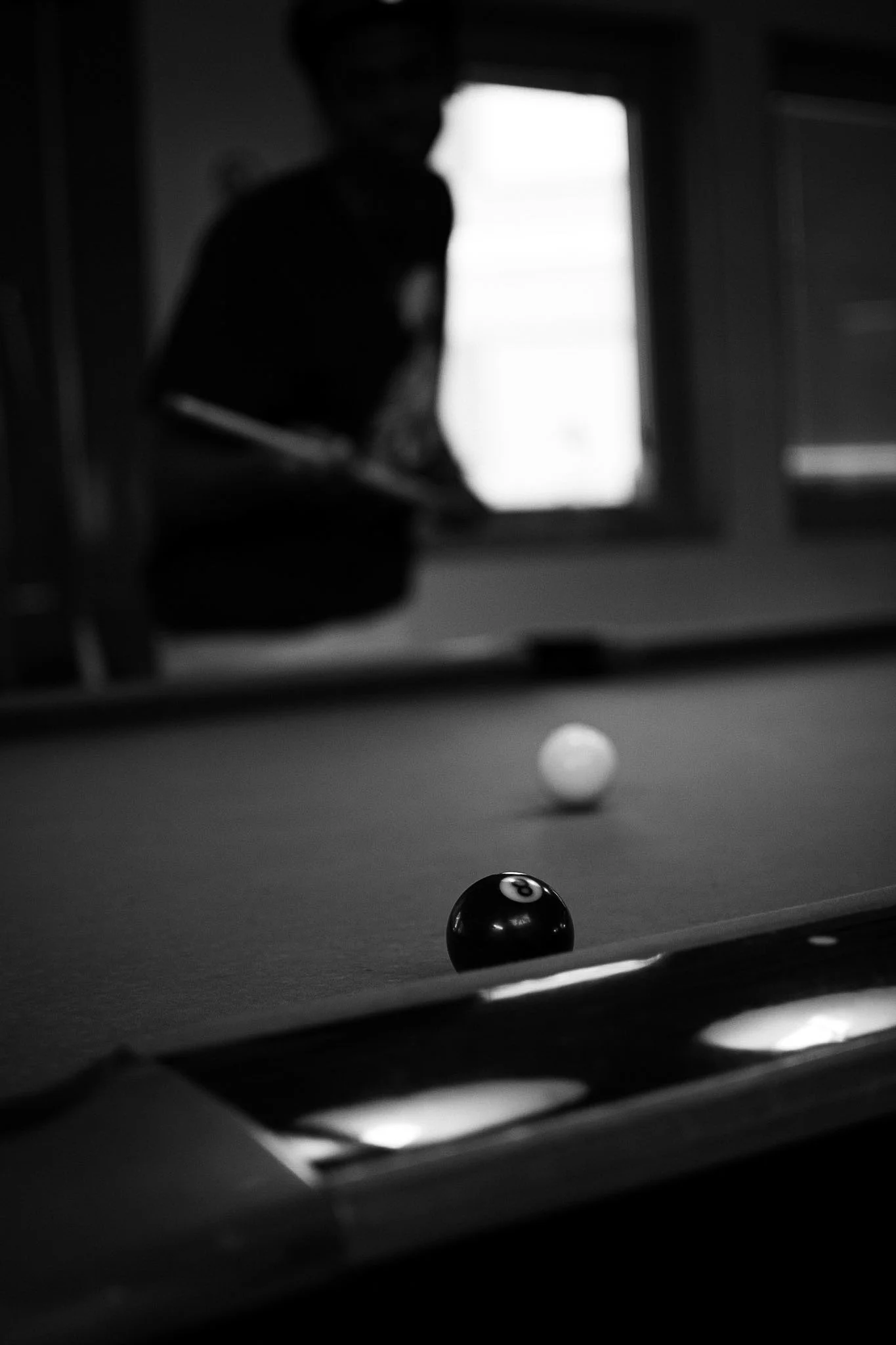 A person playing pool or billiards in a dimly lit room, with the cue ball and the 8-ball visible on the table, and another person observing in the background.