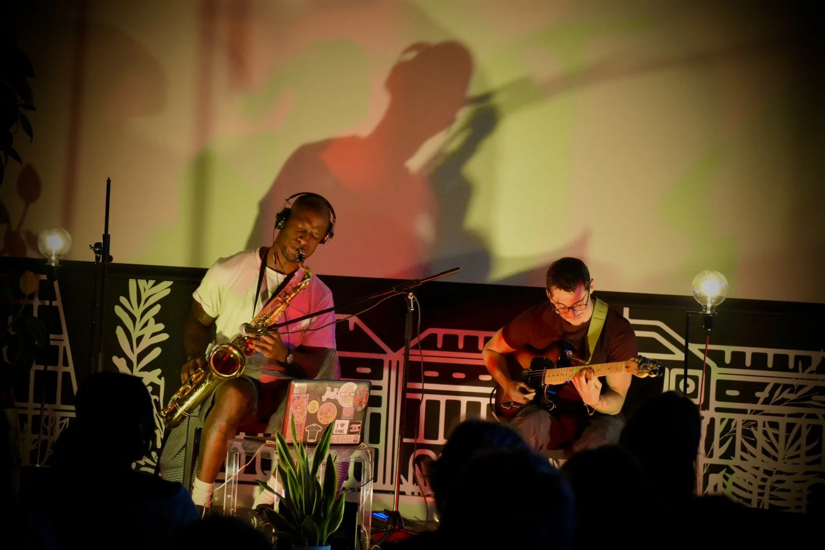 Musicians performing on stage, one playing saxophone and the other playing guitar, with a colorful backdrop and audience in the foreground.
