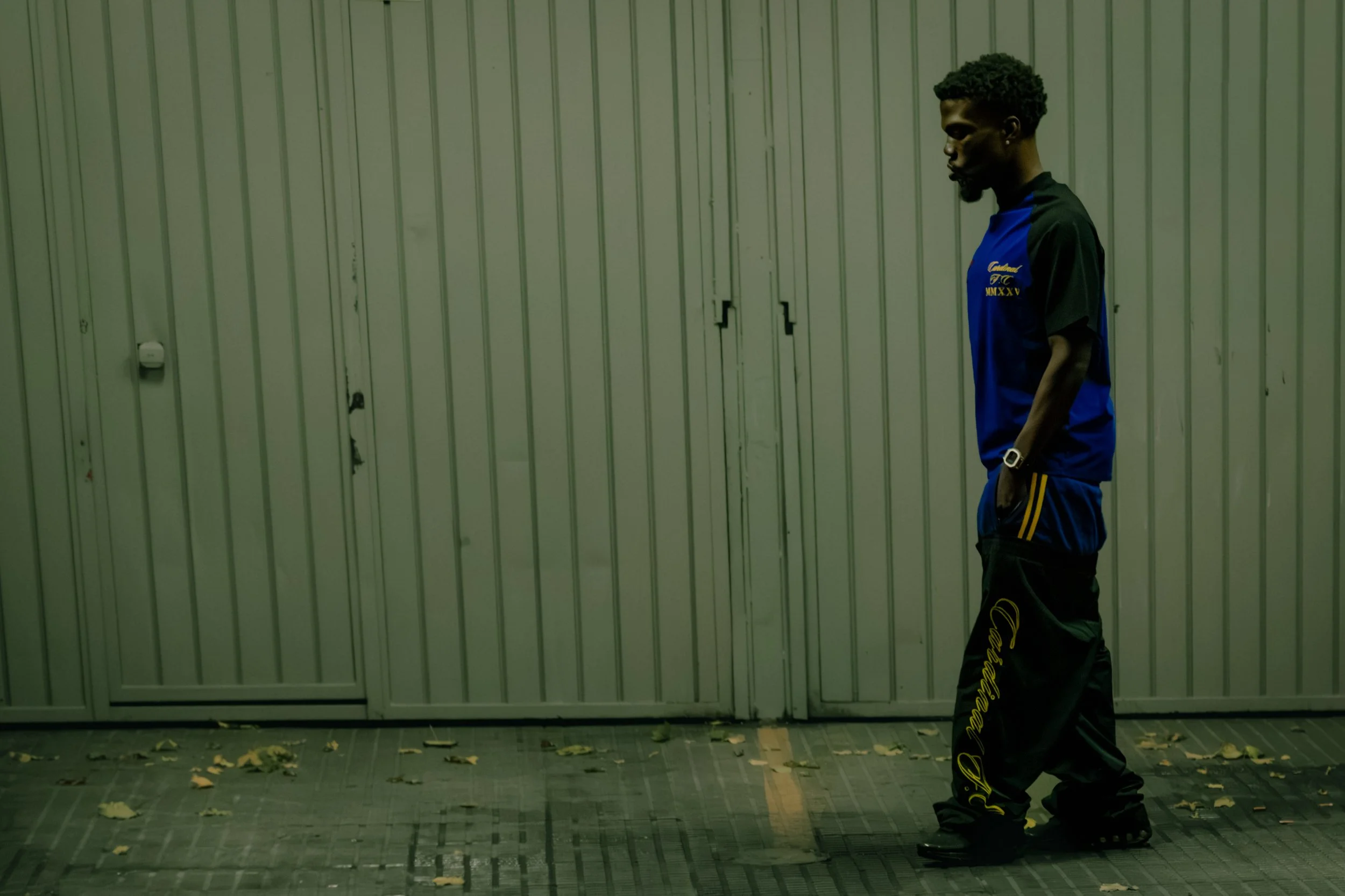 A man with dark skin, curly hair, wearing a blue and black sports outfit, and black sneakers, walks along a wall with vertical metal panels. Fallen leaves are on the ground.