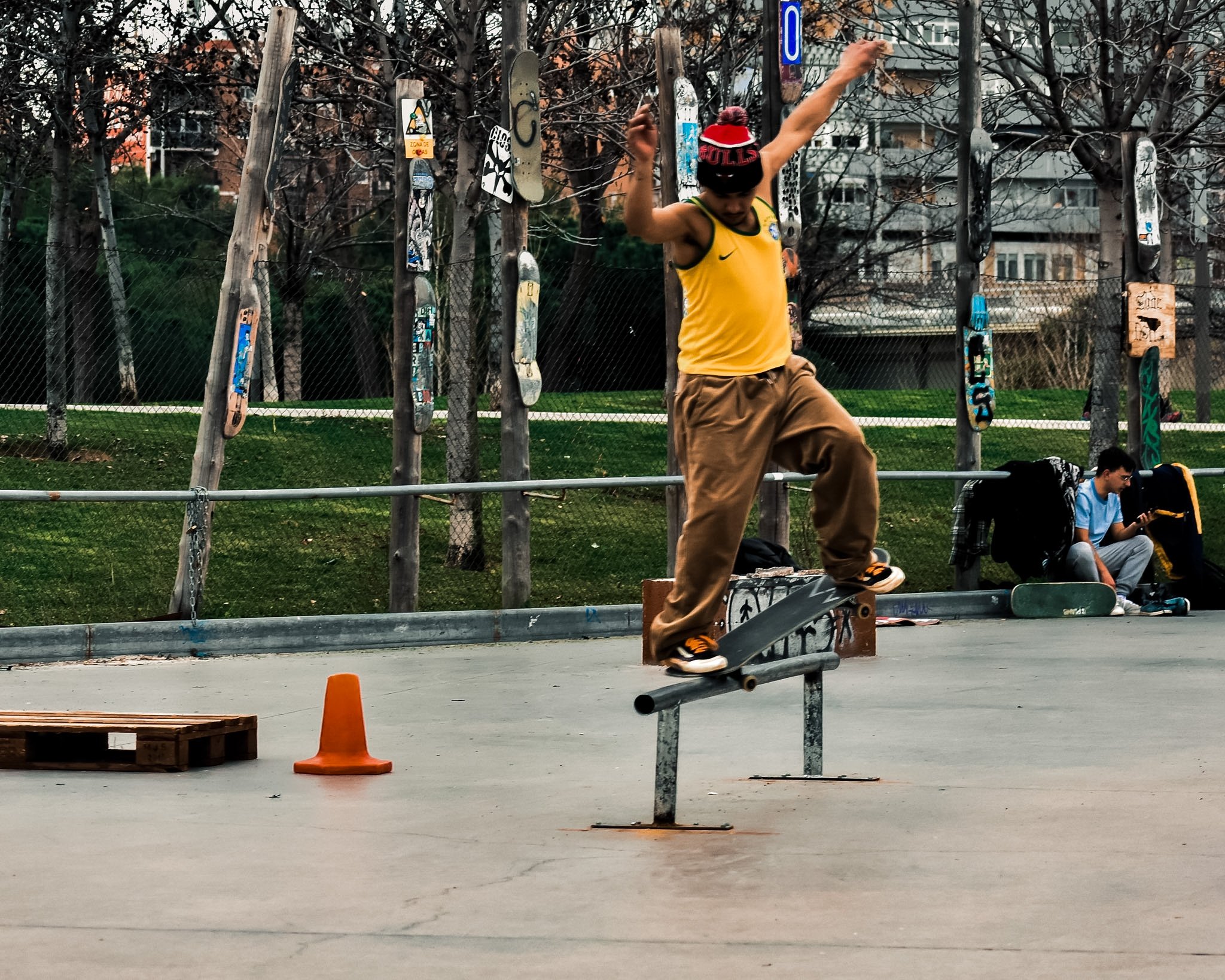 Skateboarder wearing a red hat and a yellow jersey performing a trick on a rail in a skate park, with a fence and trees in the background.