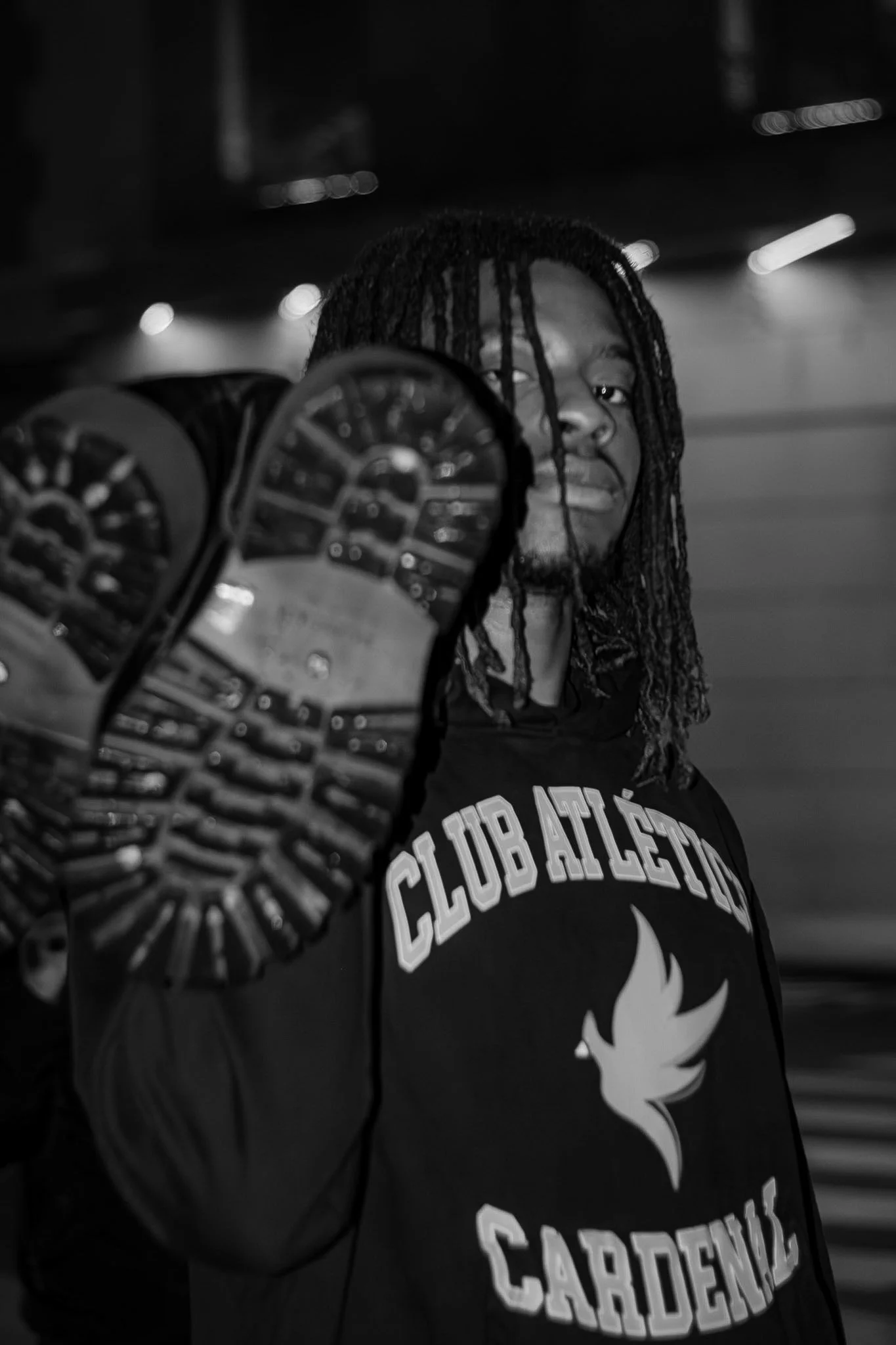 A person with dreadlocks wearing a black hoodie with white lettering and a flame graphic, holding a shoe in front of their face in a nighttime setting with blurred lights in the background.