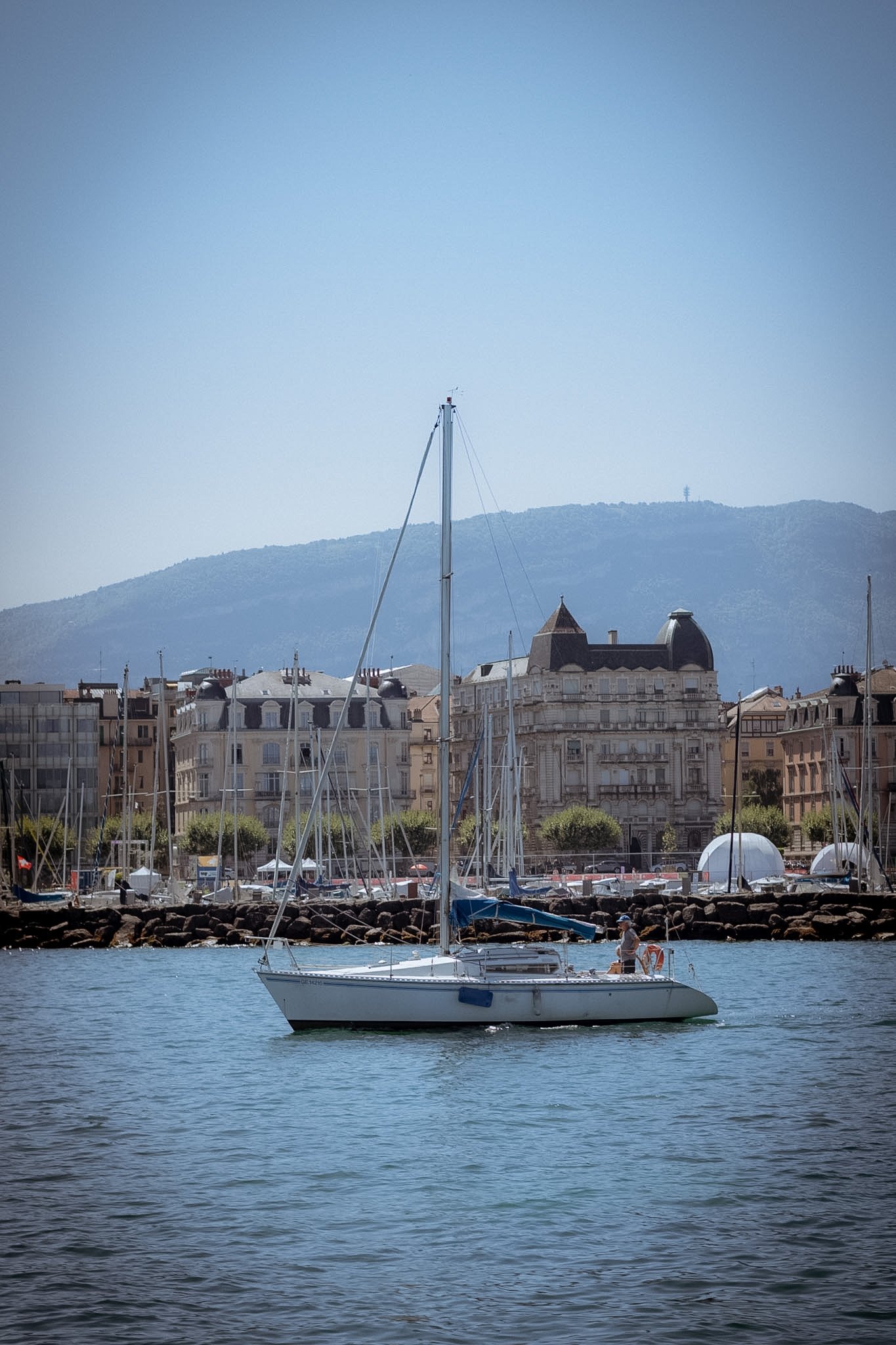 A sailboat with a person on board sailing in a harbor with buildings and mountains in the background.