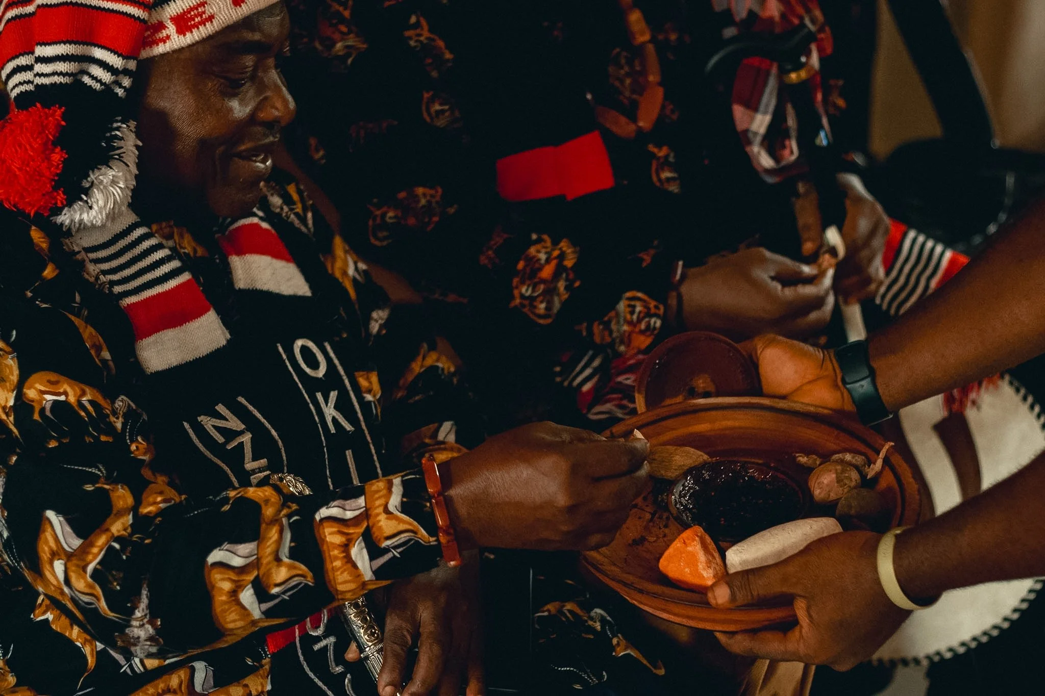 Two people wearing colorful, patterned clothing and hats, sitting closely together, possibly playing musical instruments or engaged in a cultural activity.