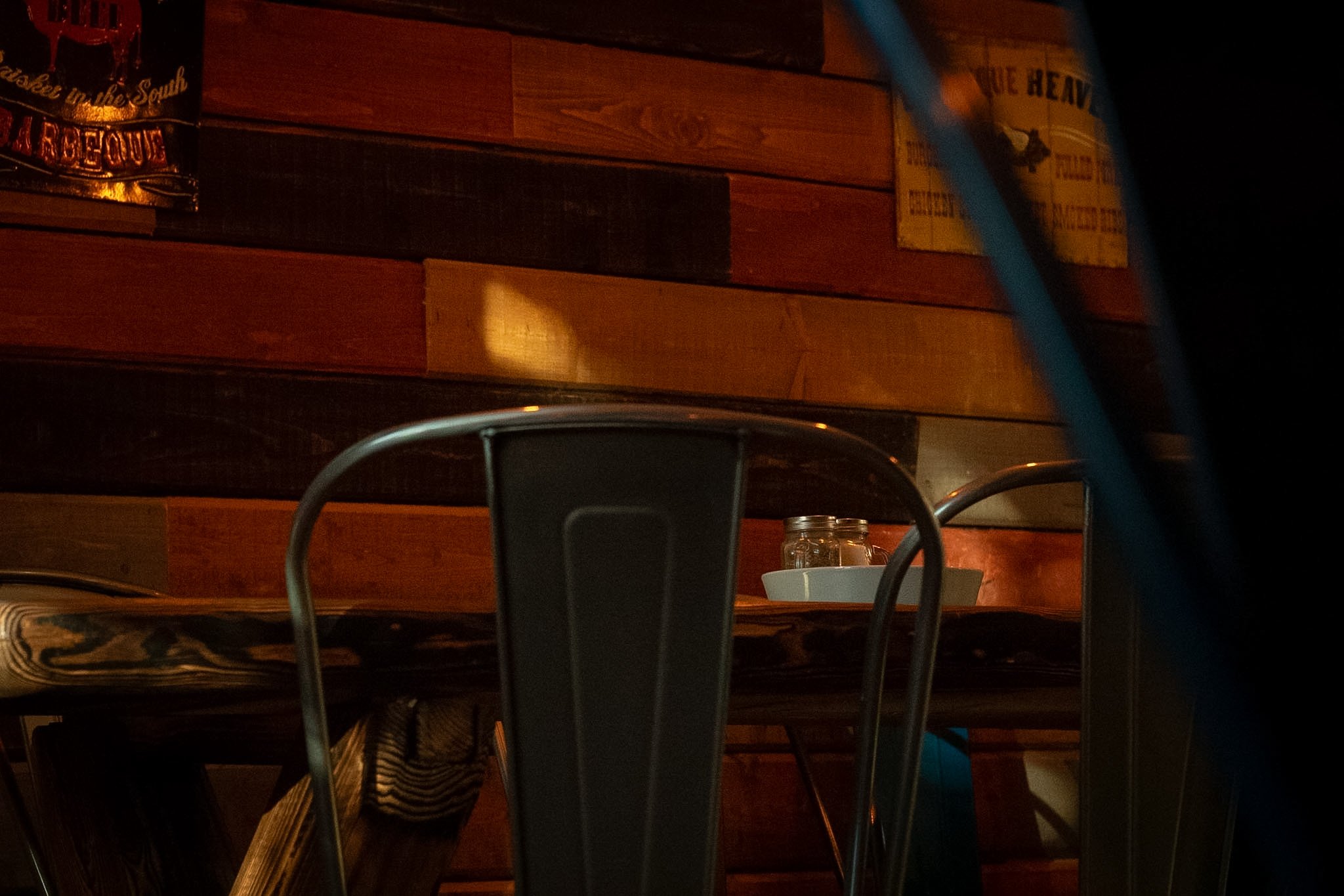A dimly lit indoor setting with a wooden wall, a metal chair, a white bowl, and a glass jar on a wooden surface, with some posters on the wall.