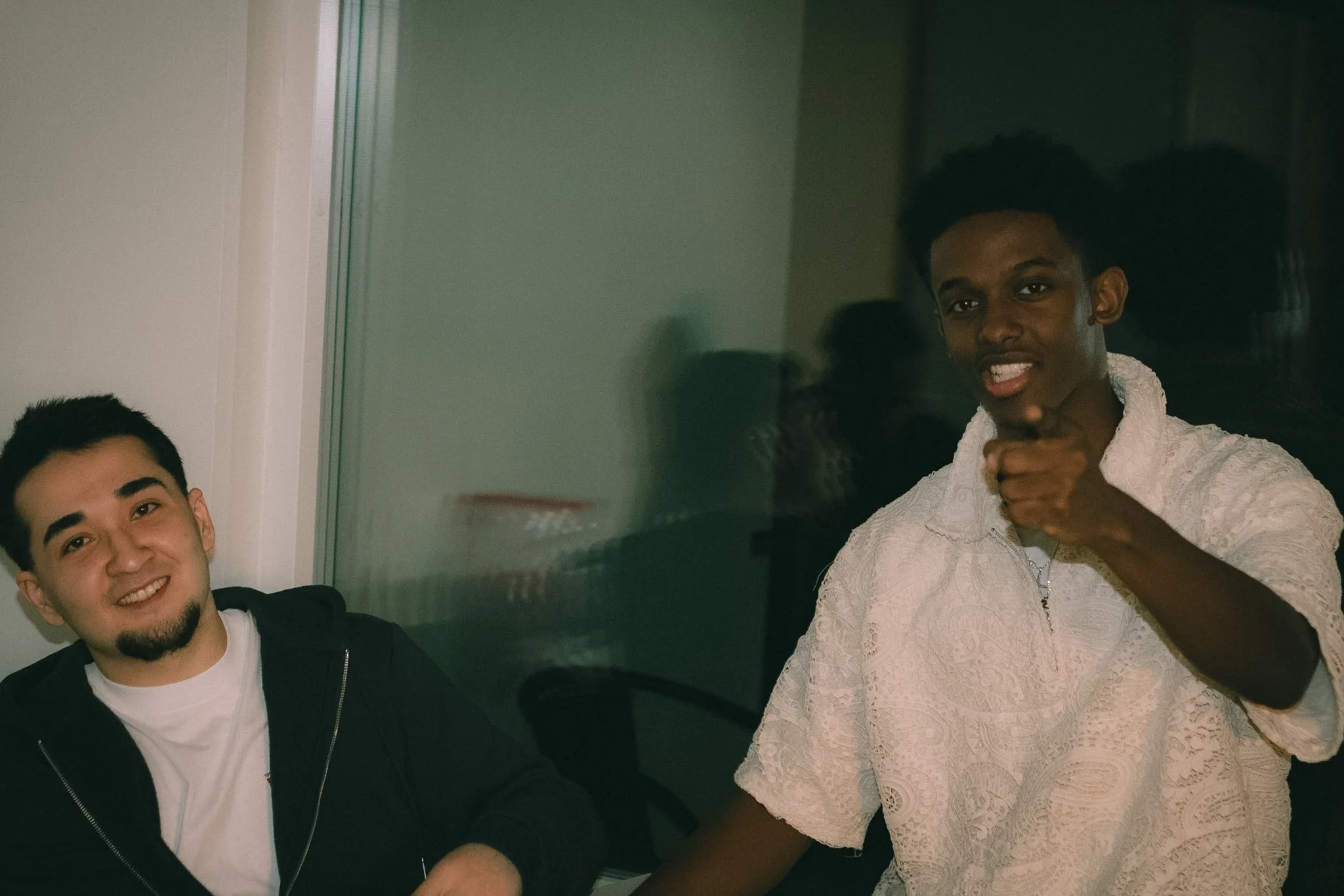 Two men are smiling and standing close together indoors. One is wearing a black jacket and white shirt, and the other is wearing a white lace top. They are reflected in a glass or mirror, with a neutral background.
