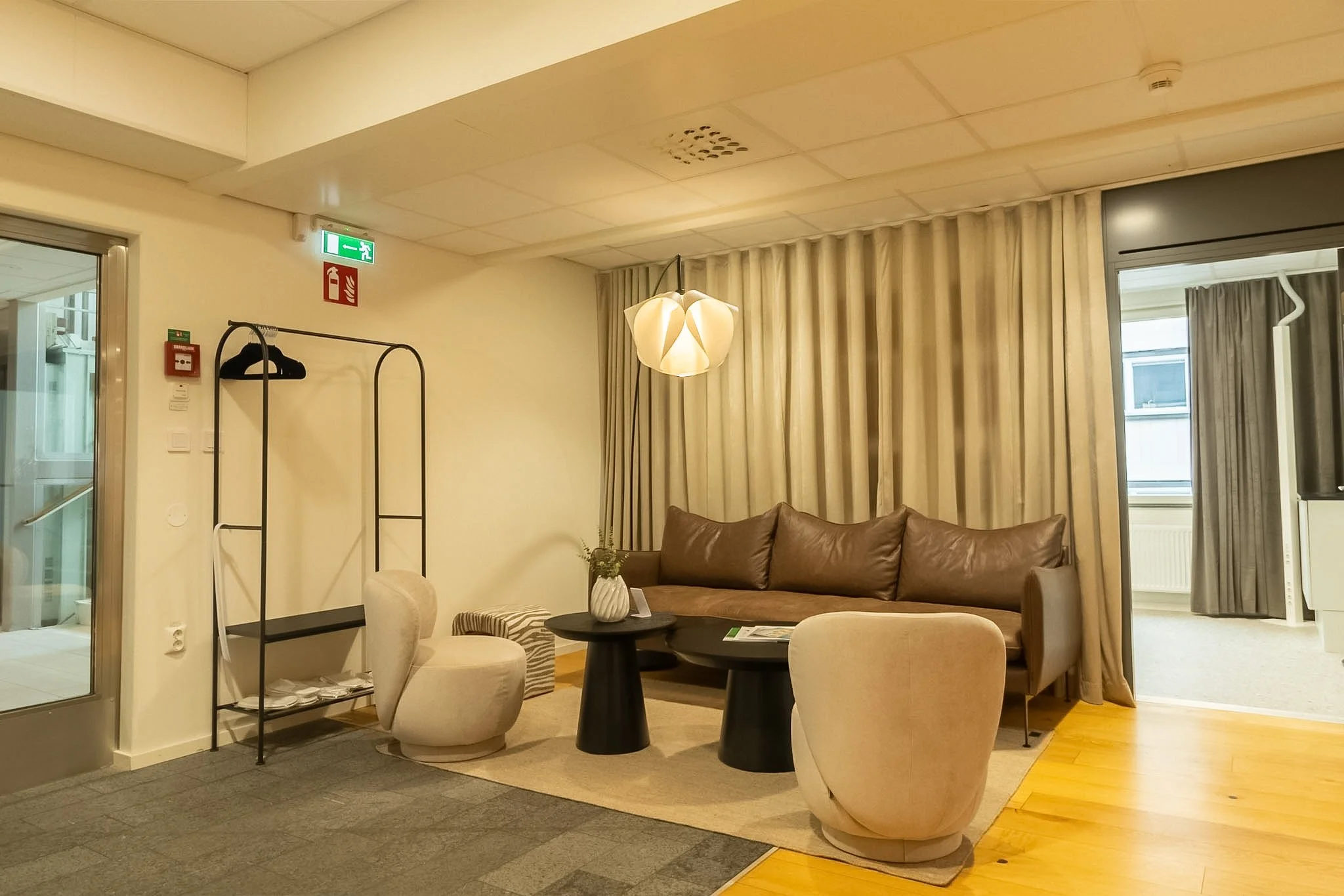 Living room with beige curtains, leather sofa, round black coffee table, accent chairs, a modern white suspended light fixture, and windows with city view. Fire extinguisher and exit sign on wall.