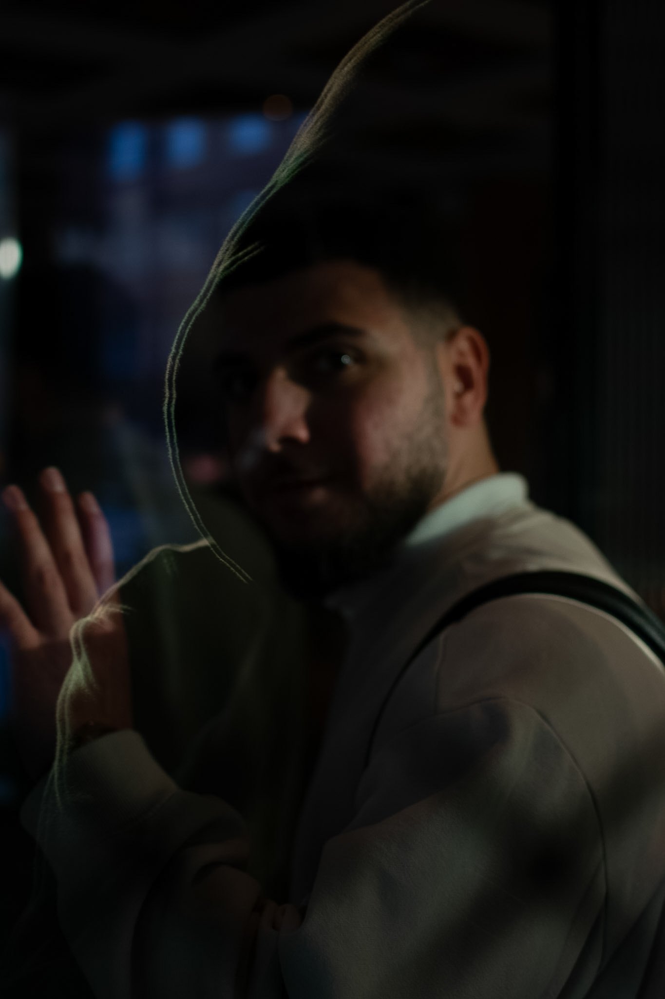 A young man in a hoodie is standing in a dark environment with a faint blue light in the background. He is looking at the camera with a slight smile and has his left hand resting against a surface.