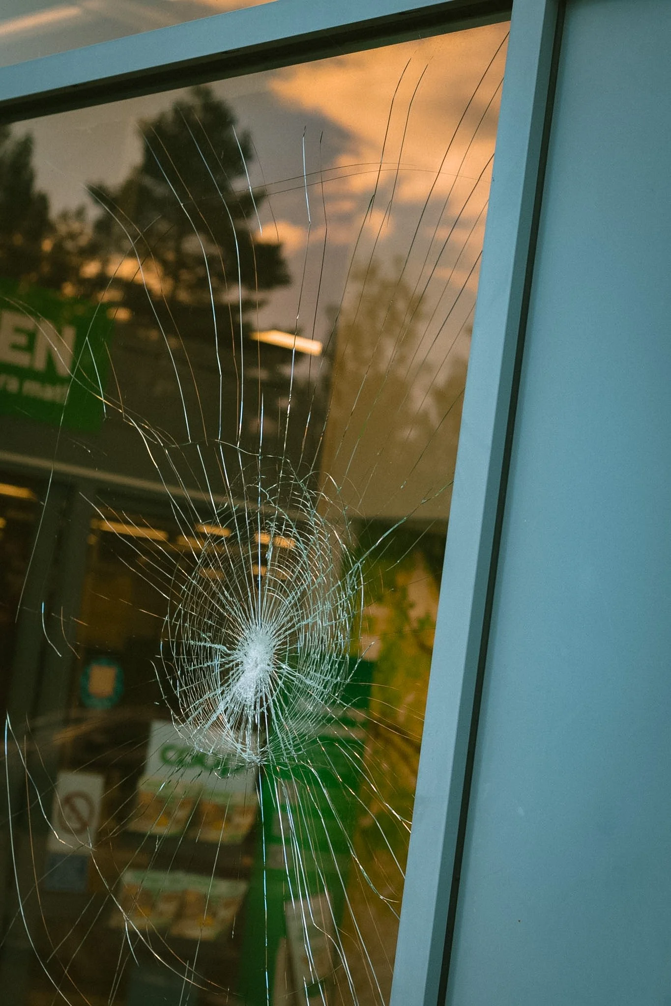 Broken glass window with a spider web fracture pattern, reflecting a sunset sky and some trees outside.