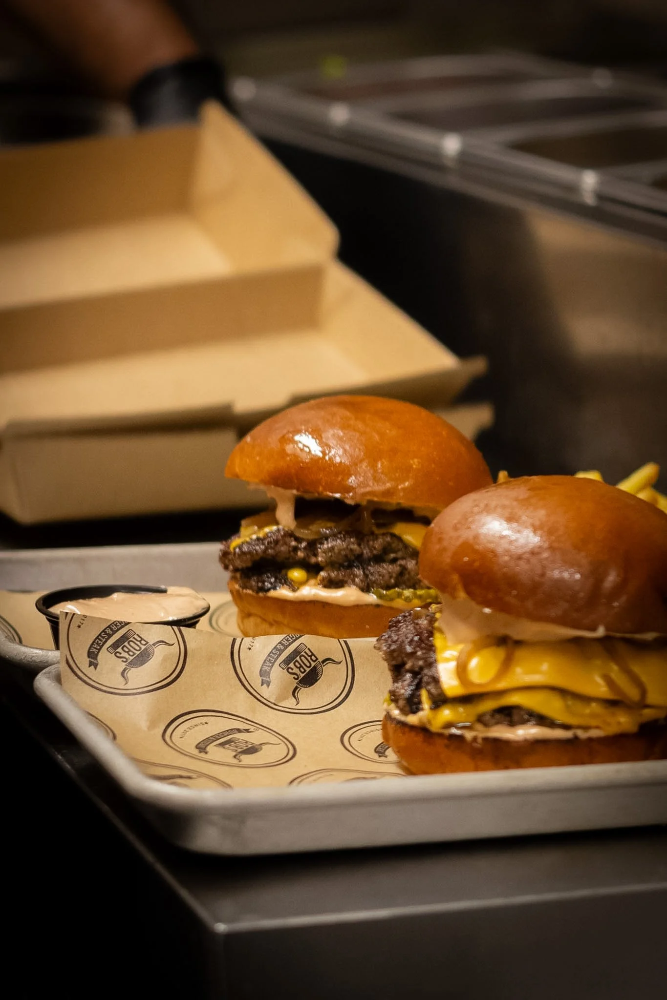 Close-up of a cheeseburger with a shiny bun, lettuce, cheese, grilled beef patty, and toppings on a paper liner with a burger logo, placed on a metal tray.