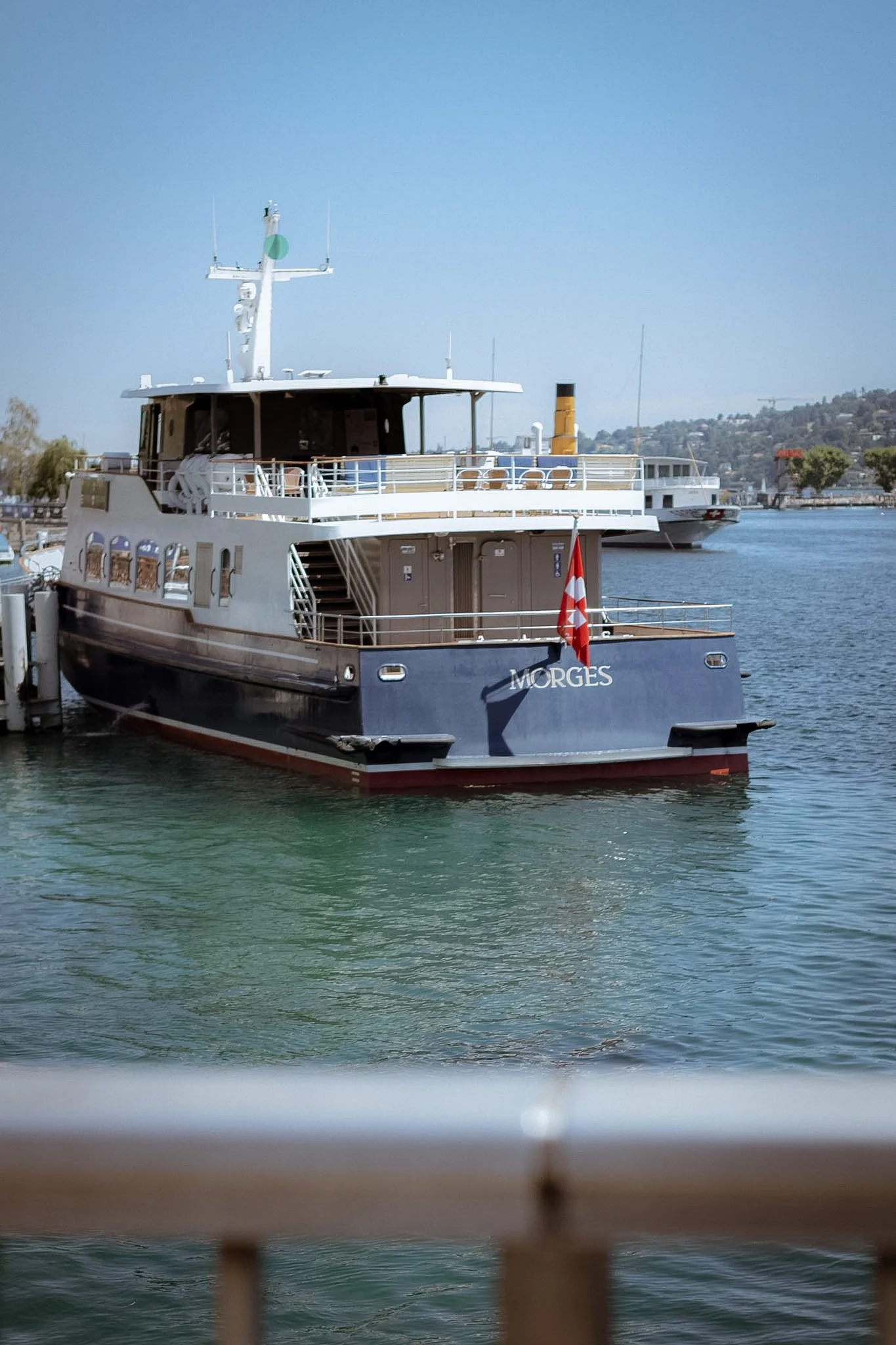 A boat named Morges docked at a marina with water in the foreground and a hillside with buildings in the background.