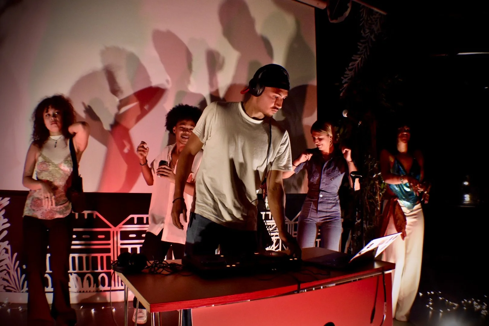 A man is DJing at a party or event with several people dancing and enjoying themselves behind him. The scene is illuminated with colorful lighting and shadows on the wall.