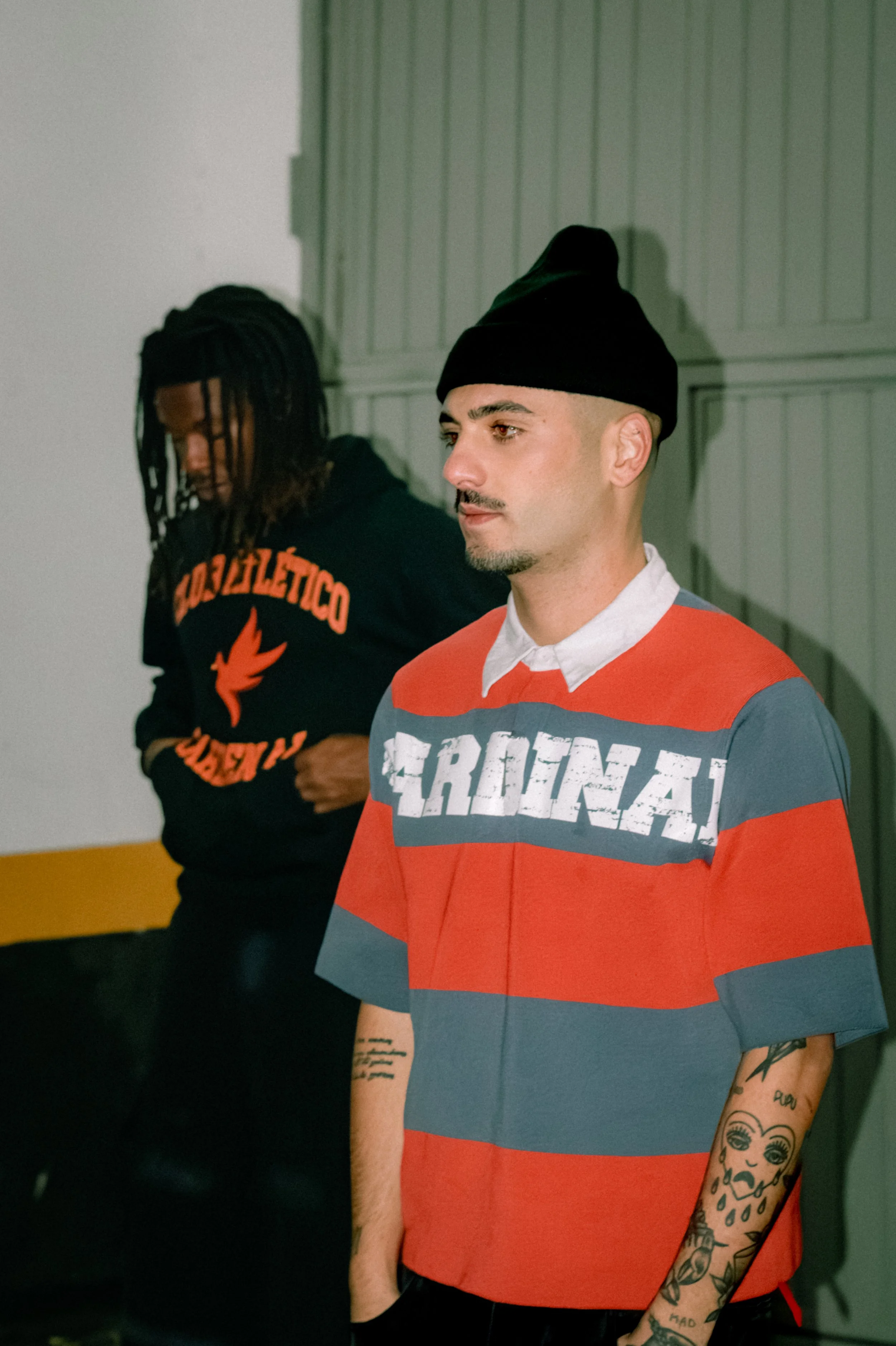 Two young men standing indoors. The man in the foreground is wearing a red and blue striped T-shirt with the word 'CROOKS' on it, a black beanie, and has tattoos on his arm. The man in the background is wearing a black hoodie with orange text and has