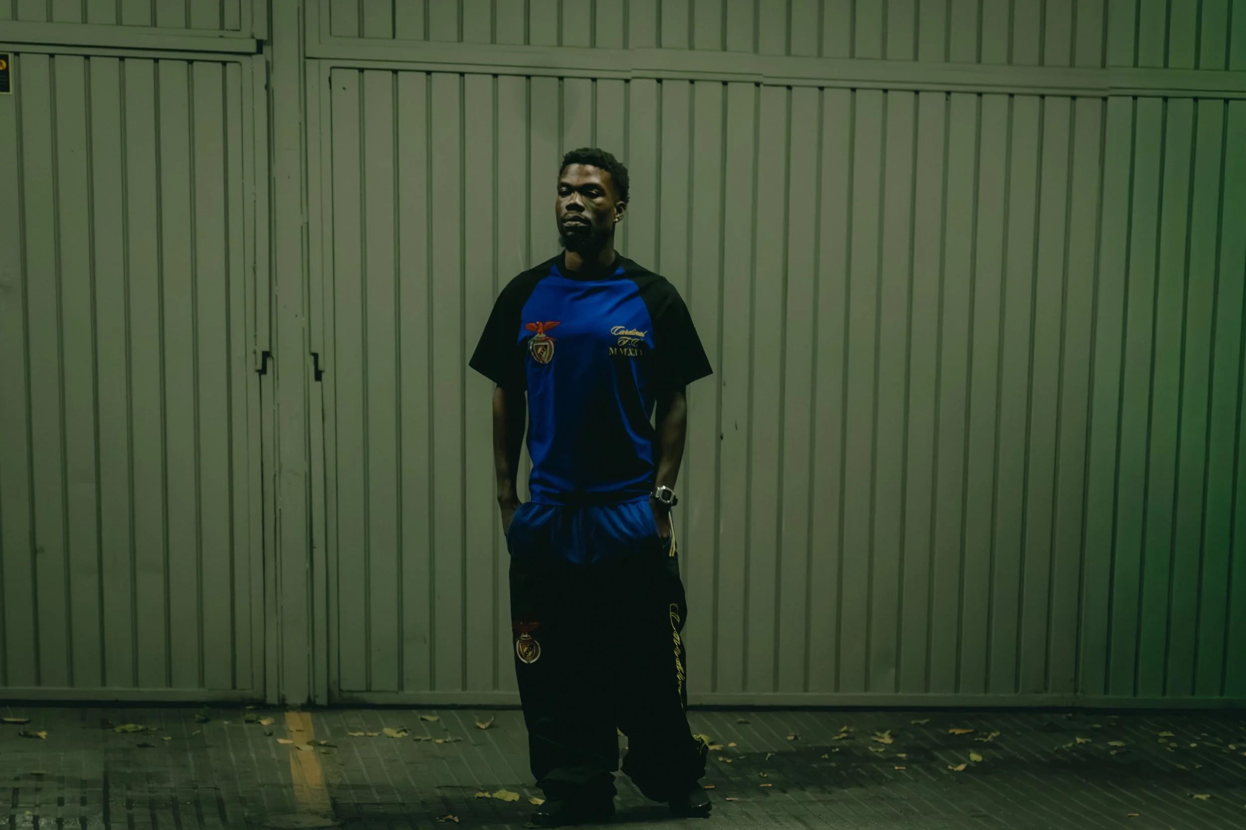A young man with dark skin, short curly hair, and a beard stands in front of a metal gate at night. He is wearing a black and blue sports shirt with logos, black shorts, and a wristwatch. He has a serious expression and his hands are in his pockets.