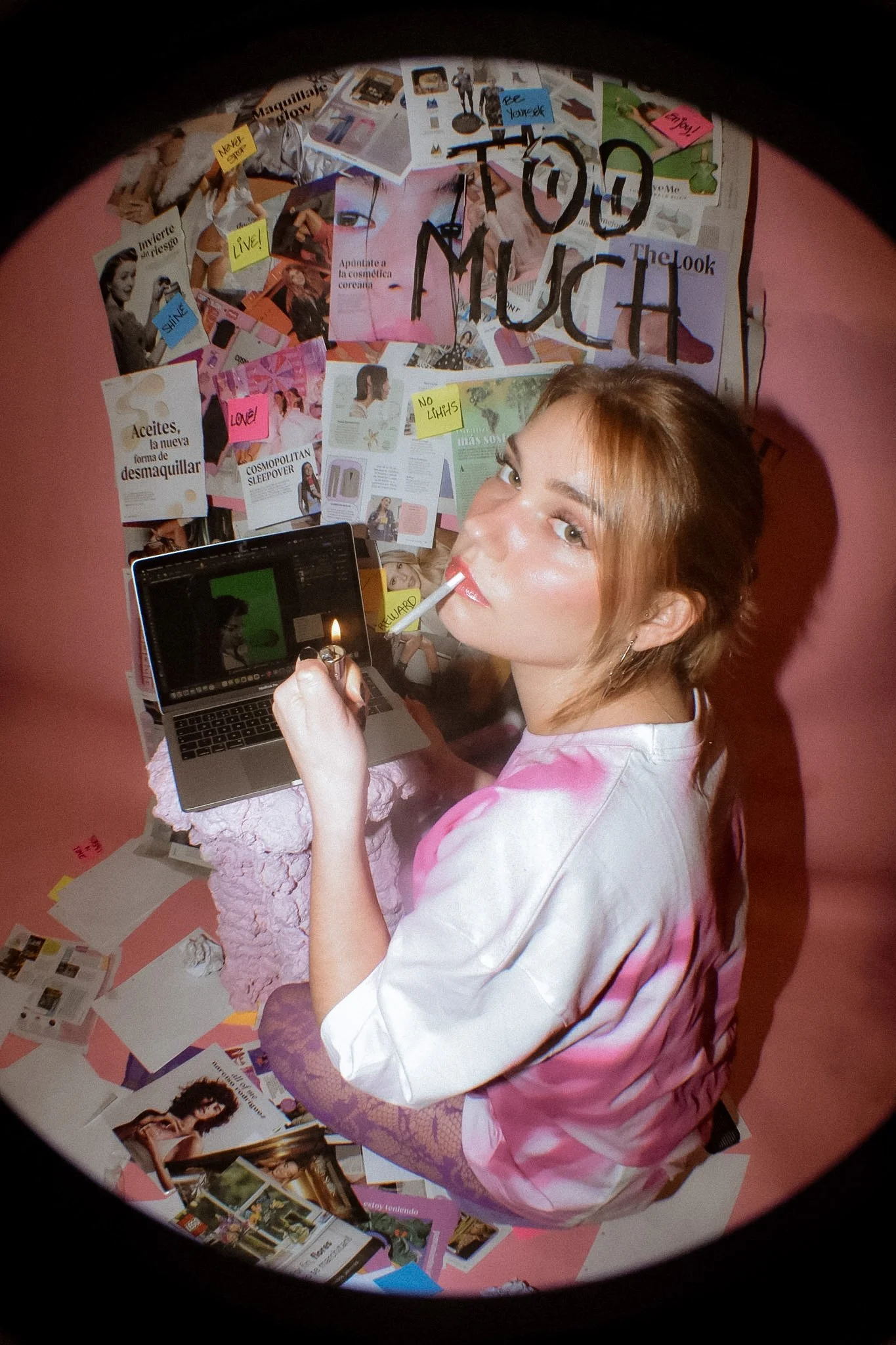 A young woman with reddish-brown hair and light skin looking up at the camera, holding a lighter. She is in front of a collage of magazine clippings on a pink wall, some of which have handwritten notes and photos.