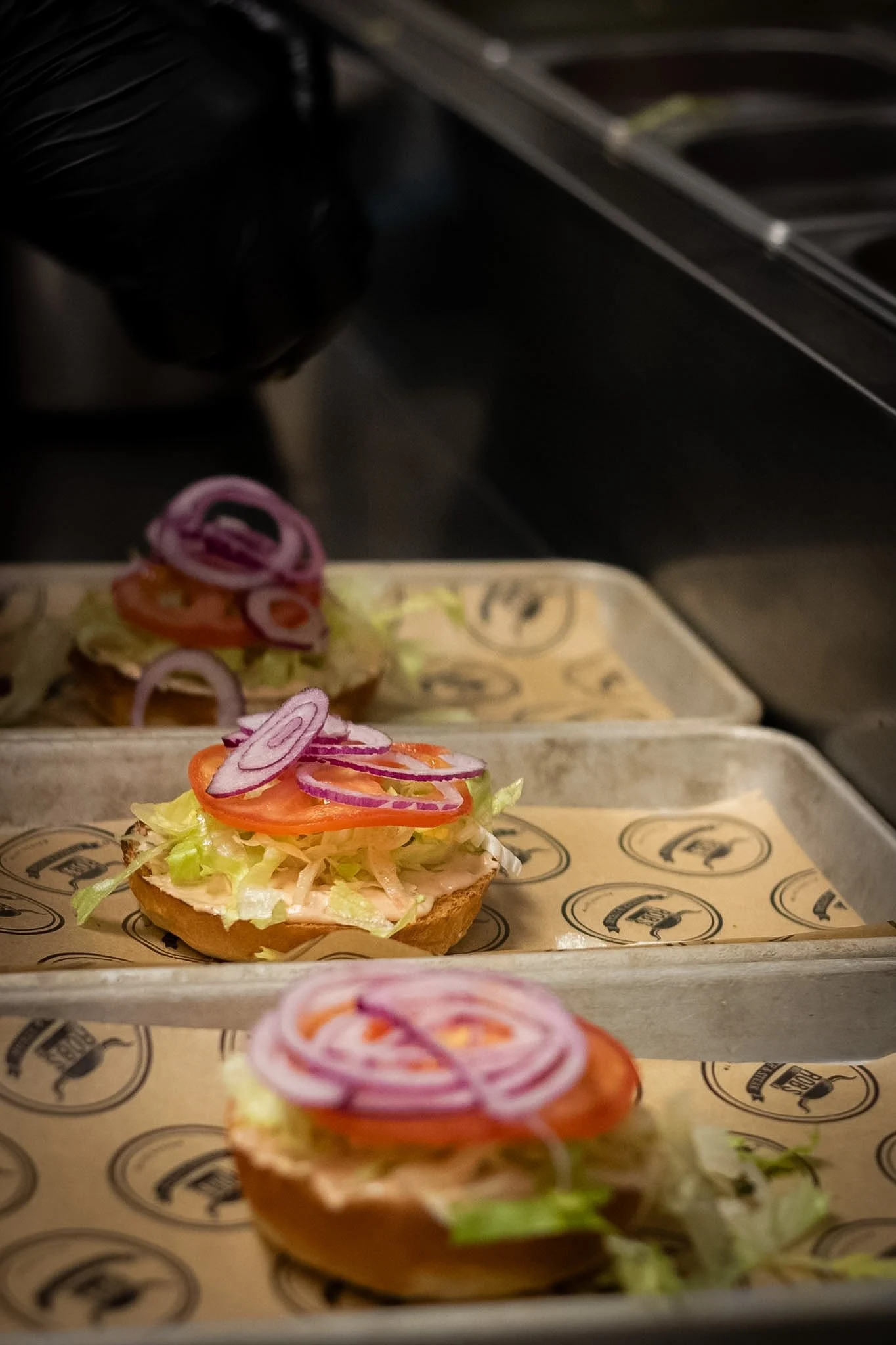 Close-up of open-faced sandwiches topped with shredded lettuce, sliced tomatoes, and sliced red onions on paper-lined trays.