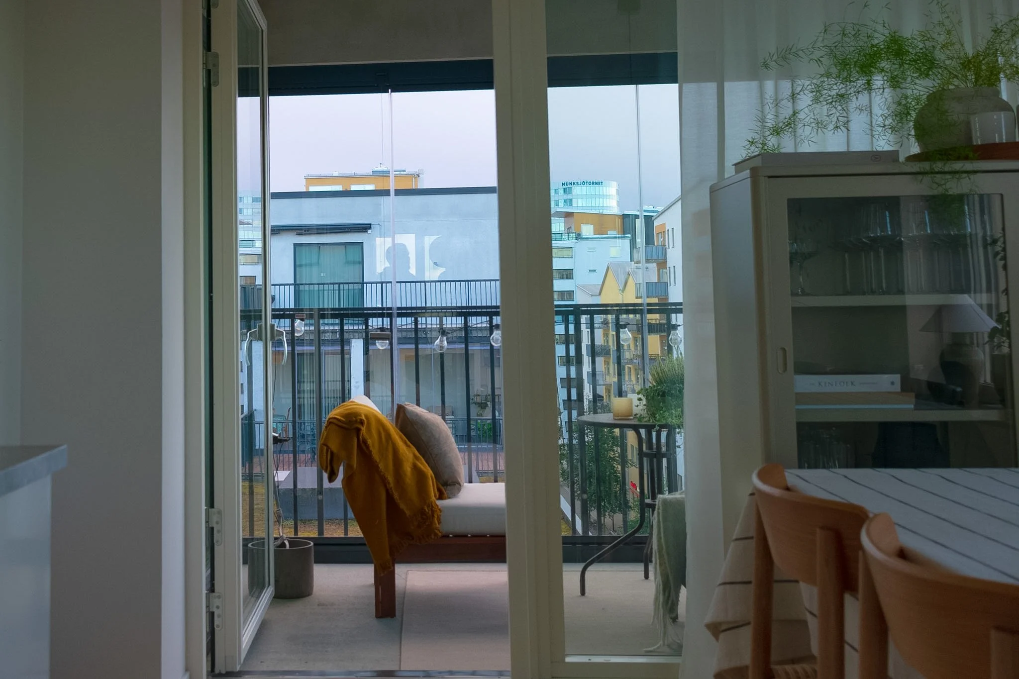 Interior view of an apartment or office with a balcony showing city buildings, glass sliding doors, a chair with an orange blanket, a small table, and a potted plant near the window.