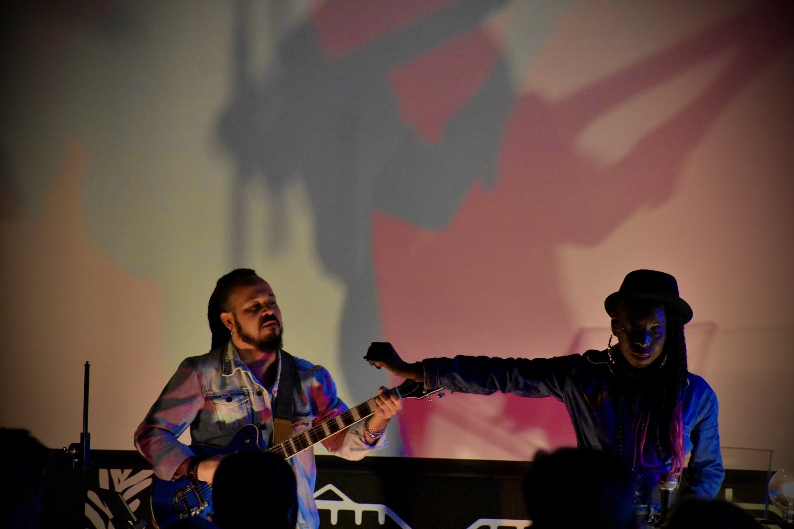 Two performers, one playing an acoustic guitar and the other extending their arm, are on stage with a large colorful projected image in the background.