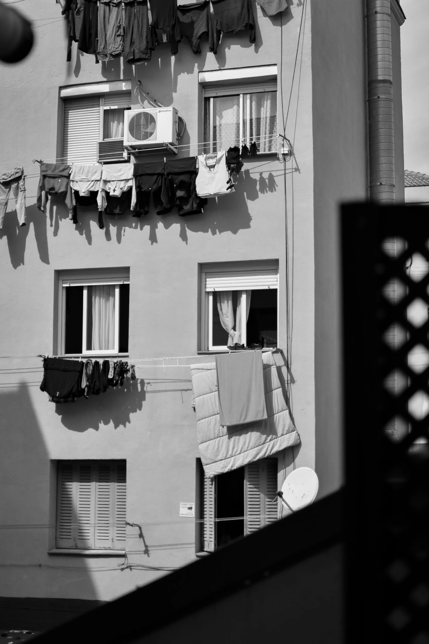 Black and white photograph of a residential apartment building with multiple windows. Clothes are hanging to dry on clotheslines outside the windows, including shirts, pants, and towels. An air conditioning unit is mounted on the exterior wall near o
