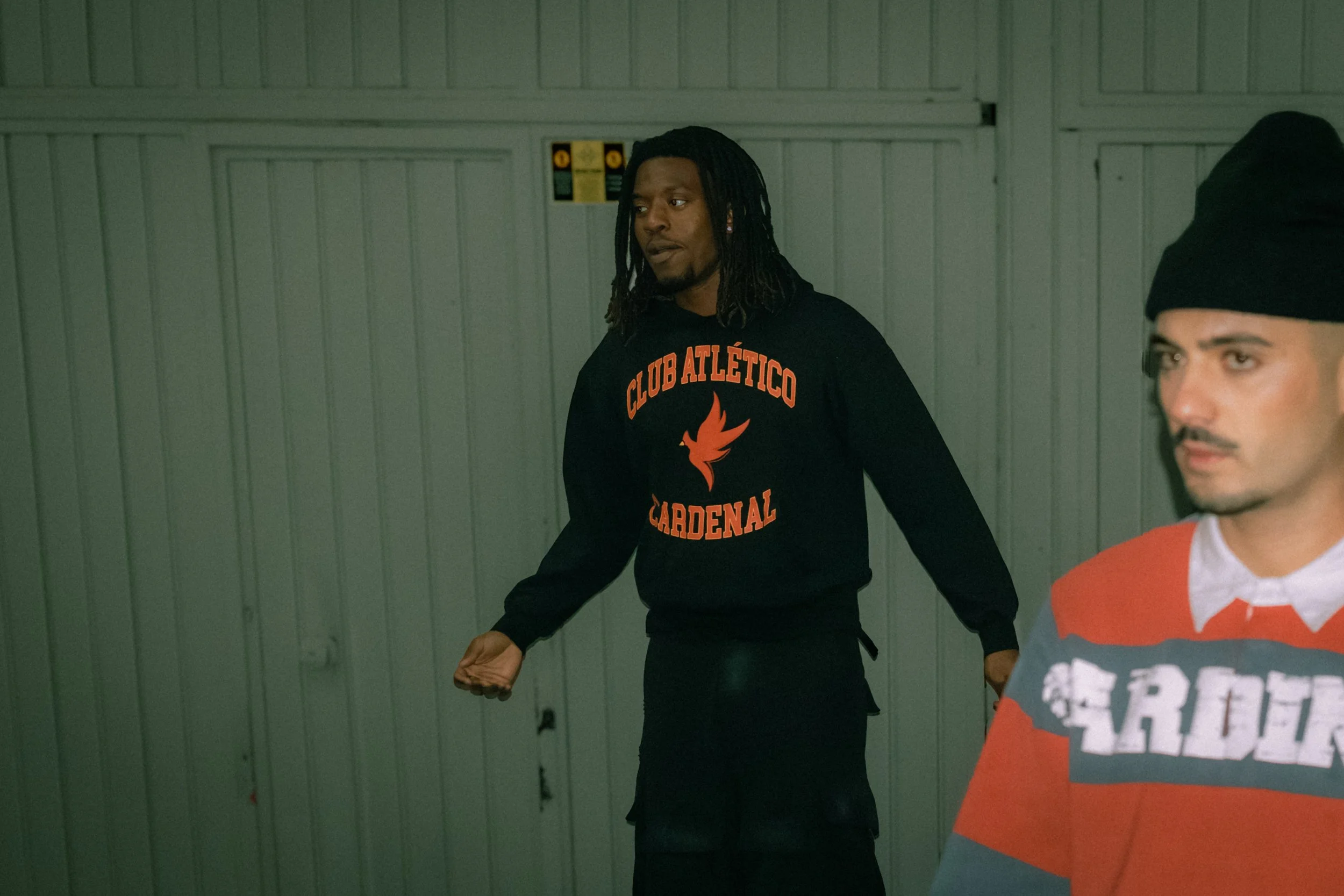 Two men standing indoors against a light green industrial wall. The man on the left has dreadlocks and wears a black sweatshirt with orange lettering and a flame logo. The other man, partially visible, has a mustache and beard, and wears a red and gr