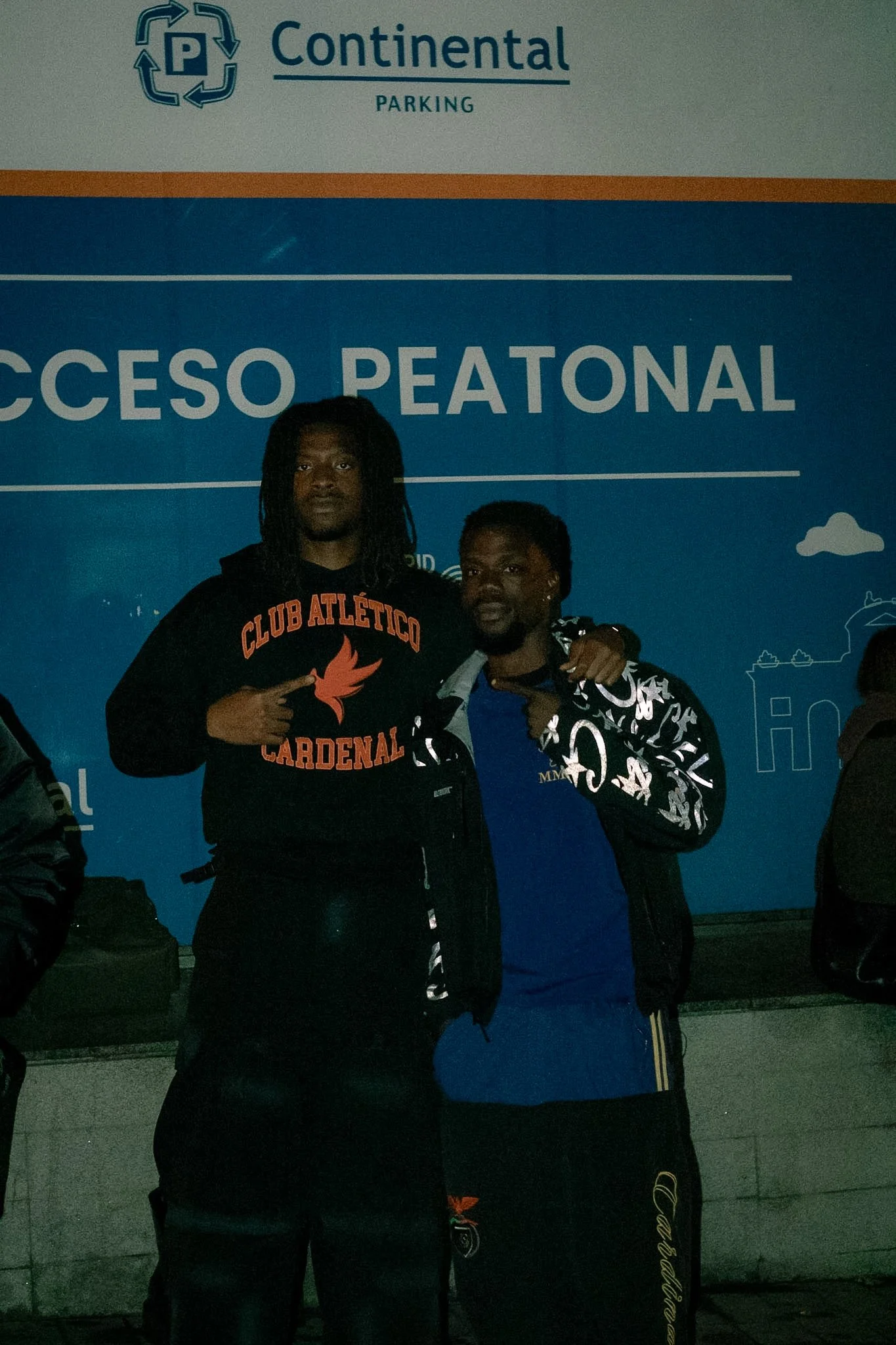 Two men standing in front of a blue sign that says 'Acceso Peatonal' with the word 'Contingent' and 'Parking' above it, in an outdoor setting at night.