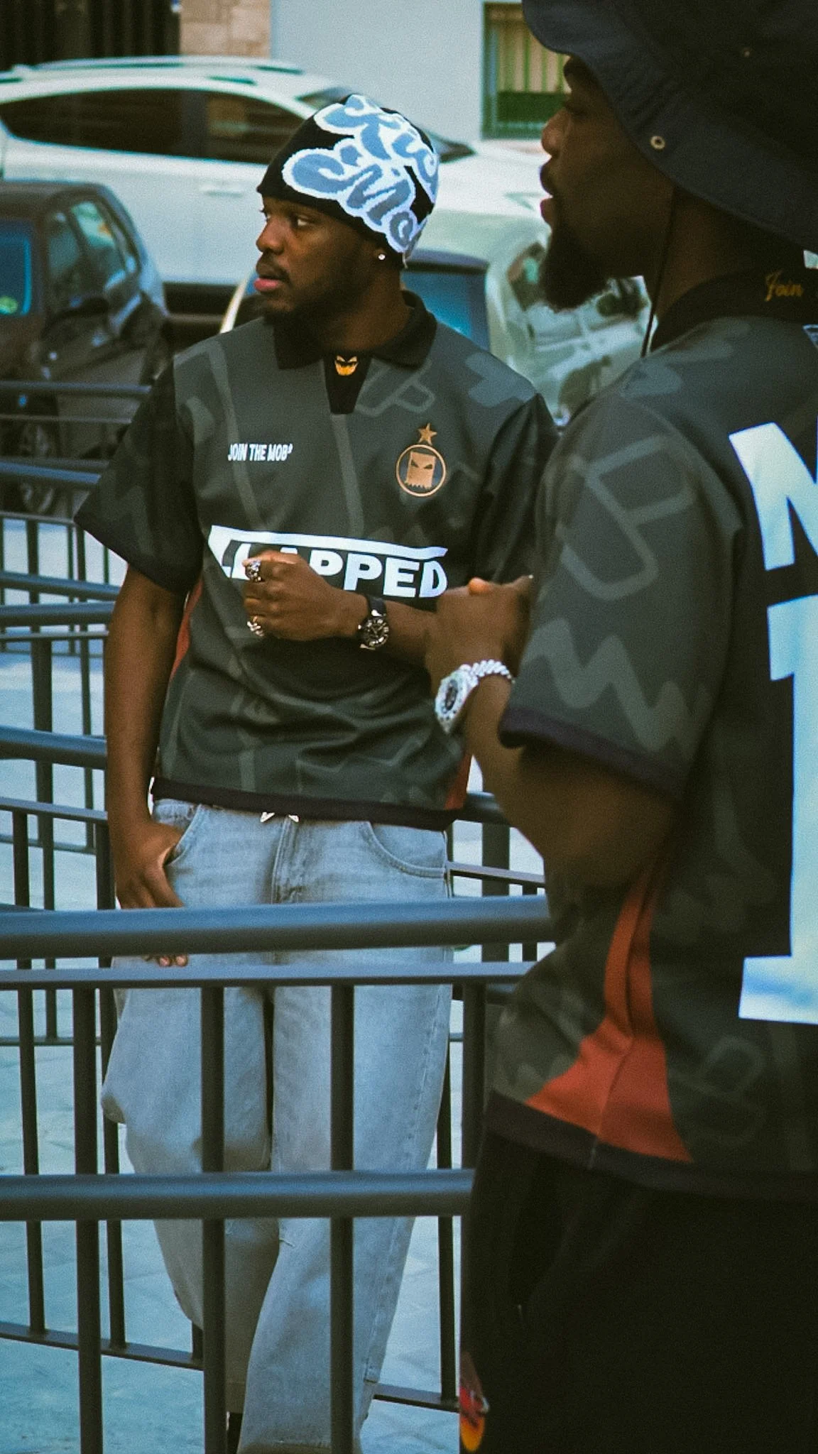 Two young men standing near a railing outdoors, engaged in conversation, one wearing a cap with white and blue patterns, the other in a black cap and t-shirt, with cars visible in the background.