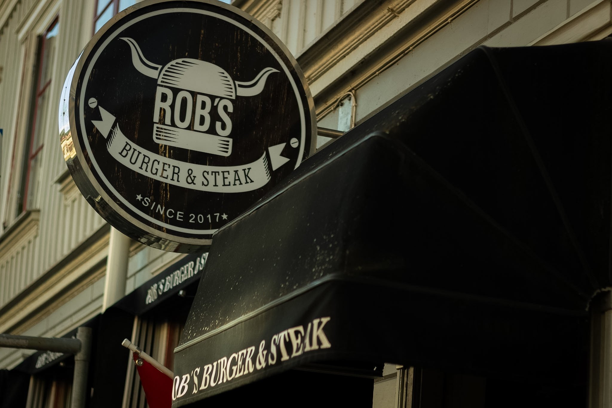 Circular black and white sign for Rob's Burger & Steak since 2017, mounted on a building exterior, featuring a stylized flame or grill graphic.