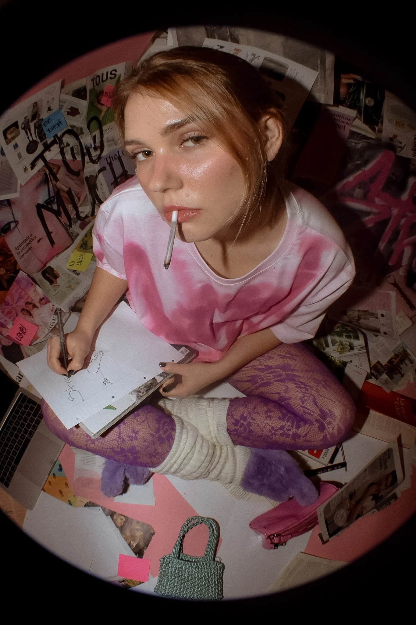 A young woman with shoulder-length brown hair, wearing a pink tie-dye shirt and purple fishnet stockings, is sitting on a colorful table. She is holding a black marker and drawing on a white sheet of paper. She has a cigarette in her mouth, and her m