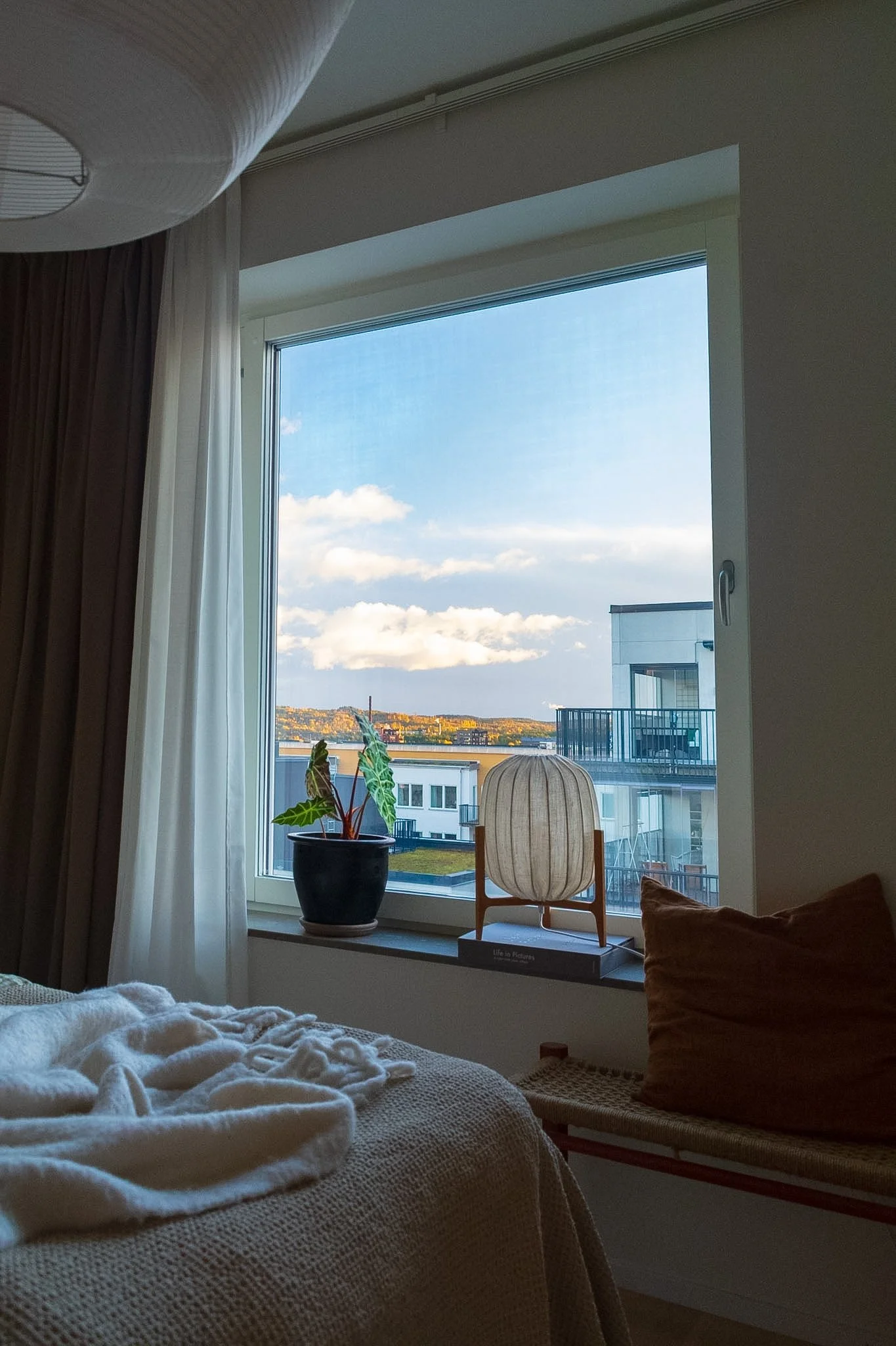 A cozy bedroom with a large window showing a cityscape and a partly cloudy sky. There is a potted plant on the window sill, a white paper lamp, and a wooden chair with a paper lantern-style shade. The bed has a white textured cover and a brown pillow