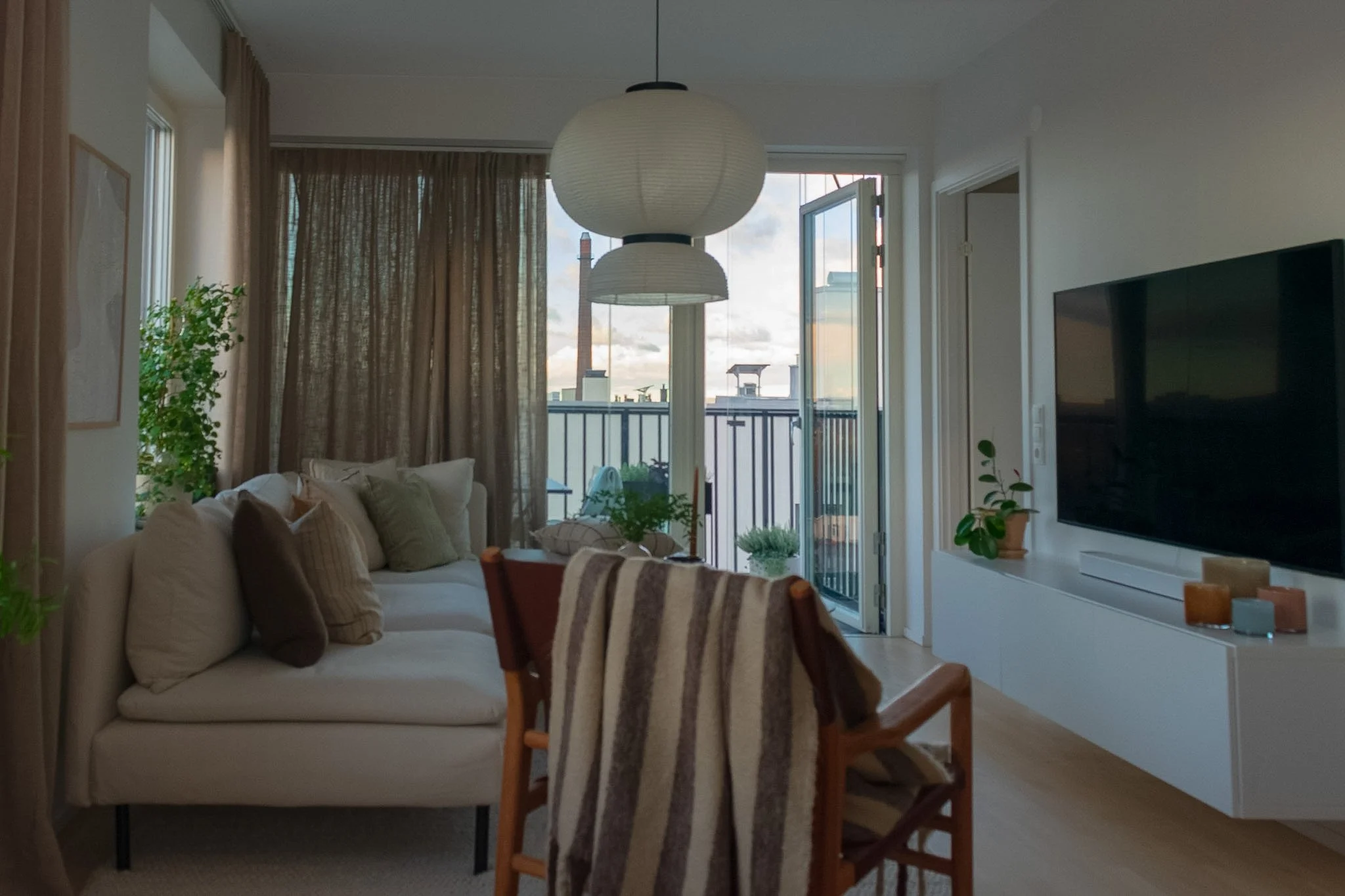 Living room with beige sofa, wooden dining chairs with striped cushions, potted plants, a large flat-screen TV, glass door leading to balcony, and round paper lantern ceiling lights.