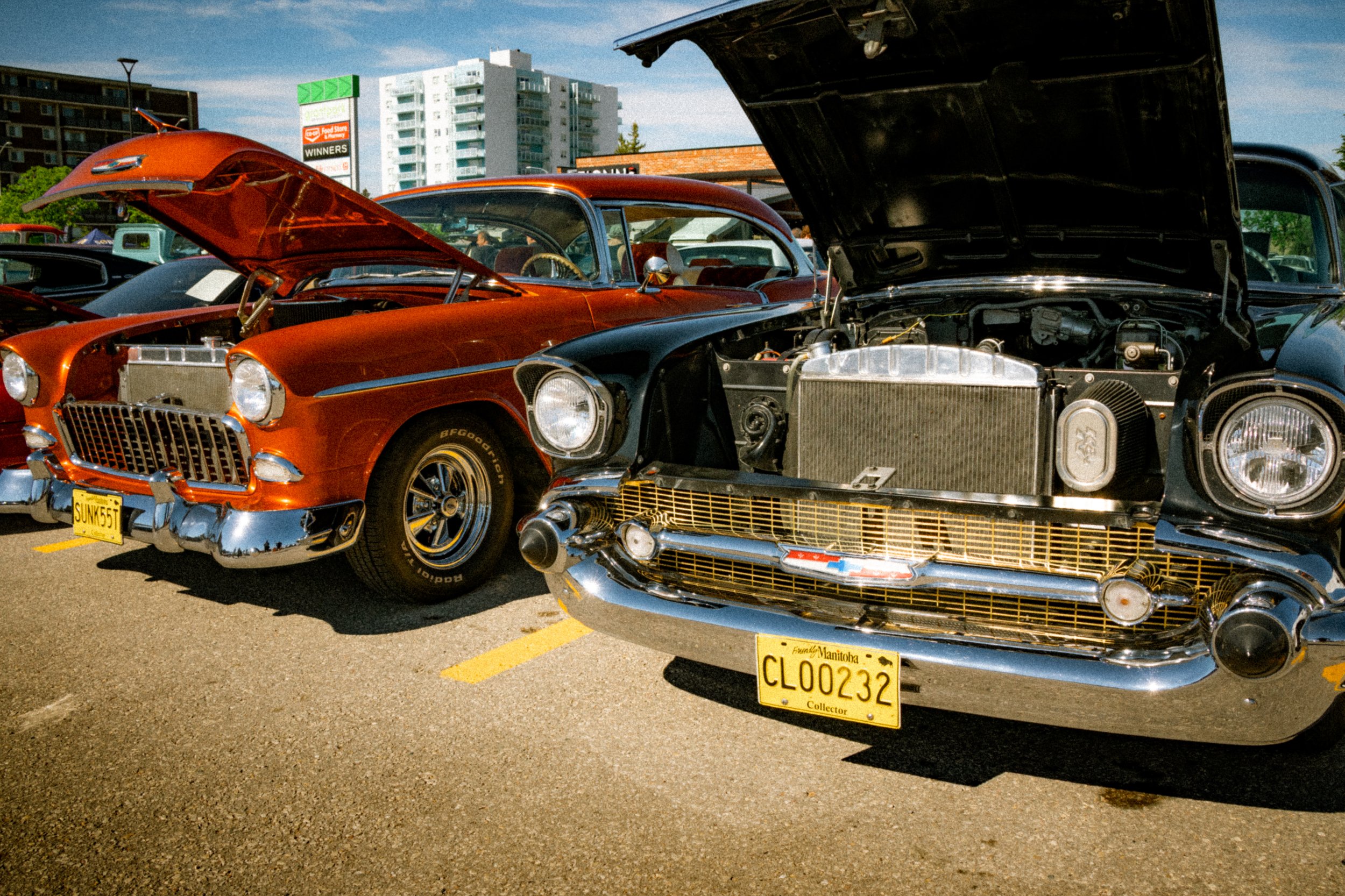 Several vintage cars parked at an outdoor car show with their hoods open, including a bright orange classic coupe and a black car with a chrome grille. Background shows a cityscape with tall buildings and clear skies.
