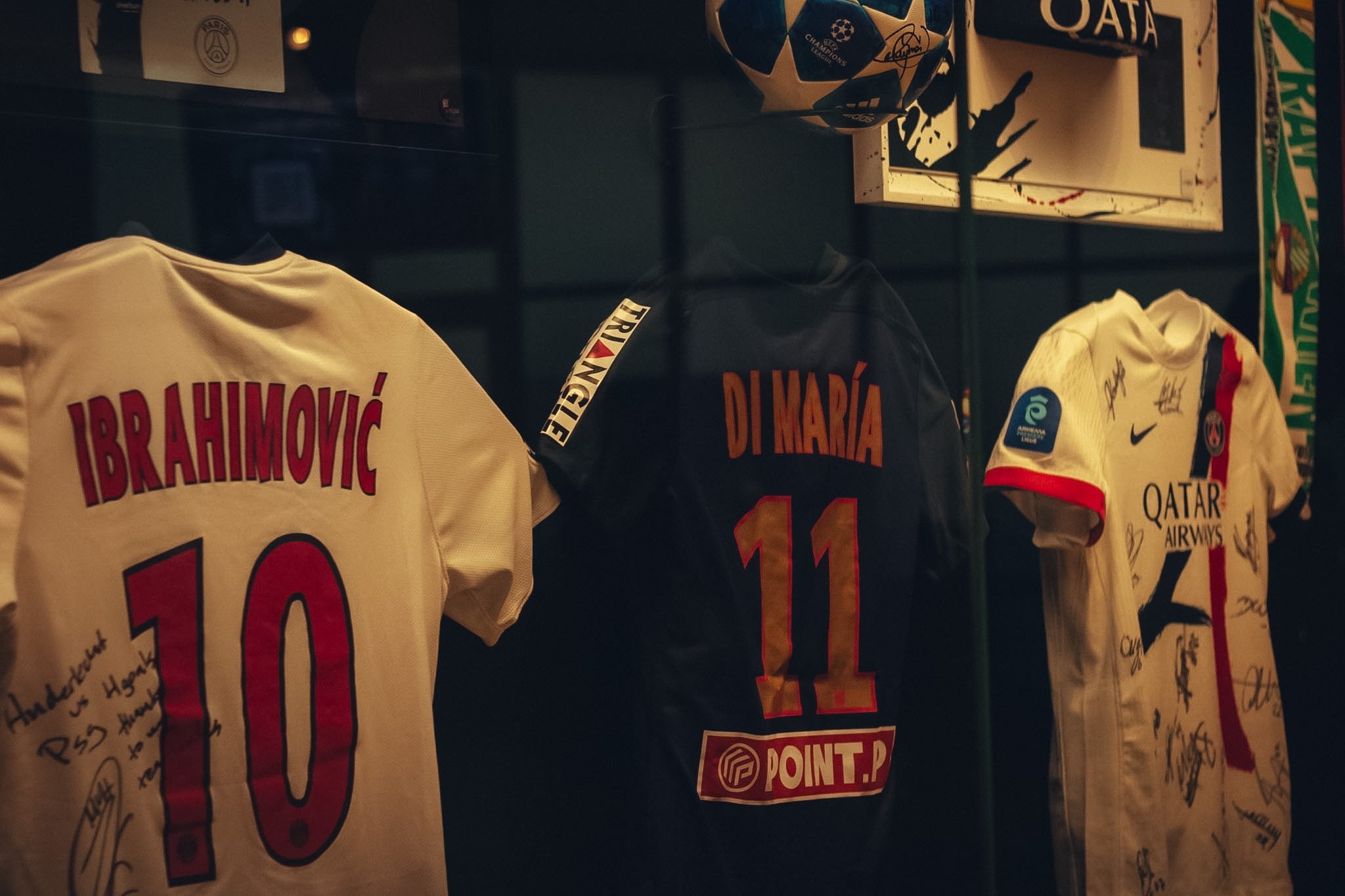 Display of signed soccer jerseys with signatures, including players' names like 'Brahimović' and 'Di Maria', and a signed soccer ball on a shelf.