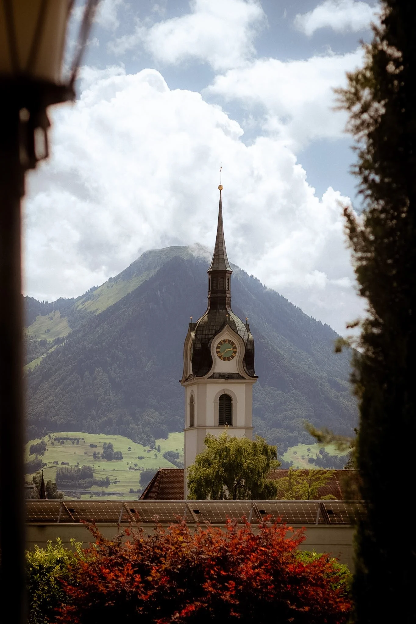 A church steeple with a clock against a mountainous backdrop with clouds in the sky, framed by trees and a garden in the foreground.