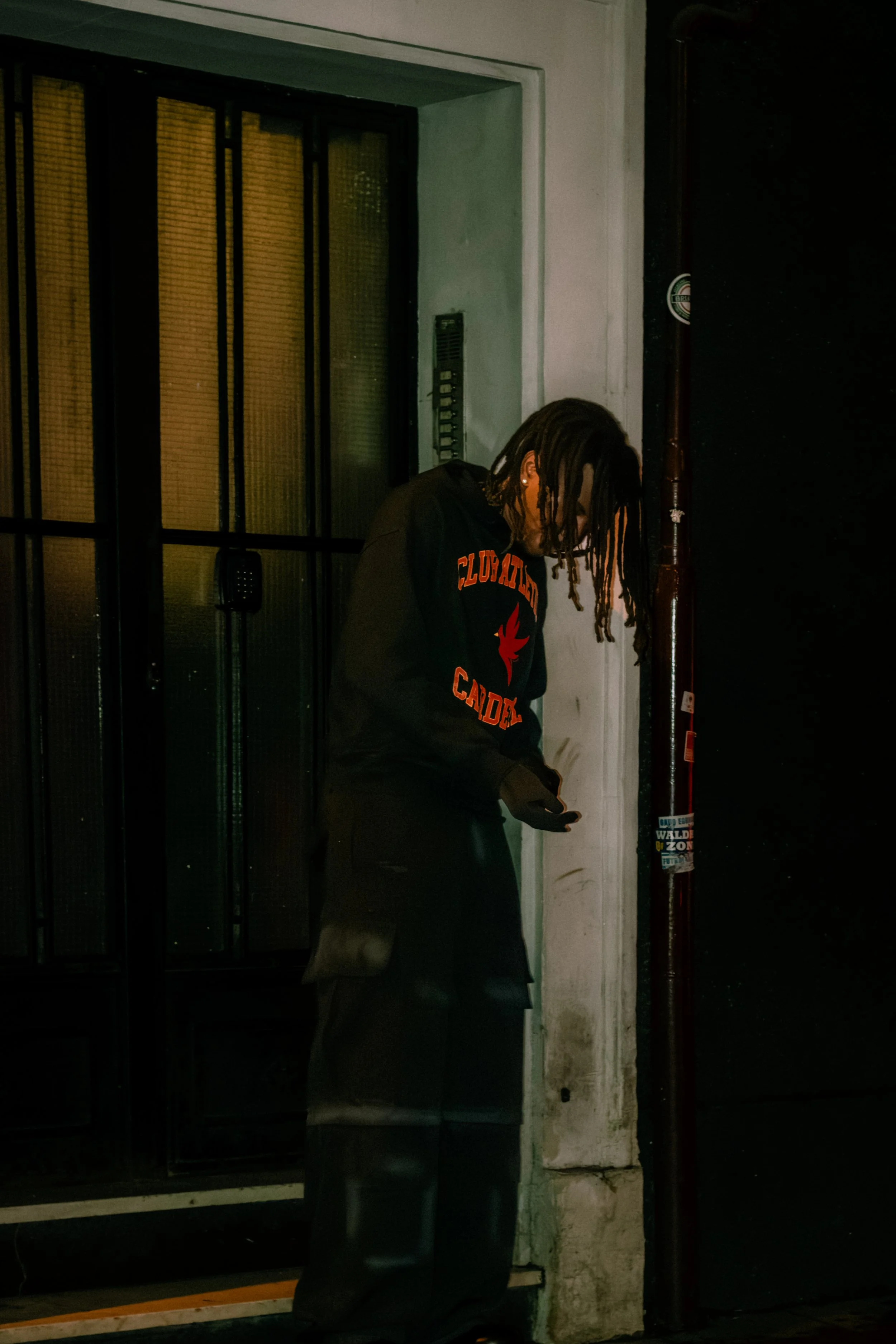 A person with dreadlocks wearing a black hoodie with orange text, standing in front of a building at night, looking down at their phone.