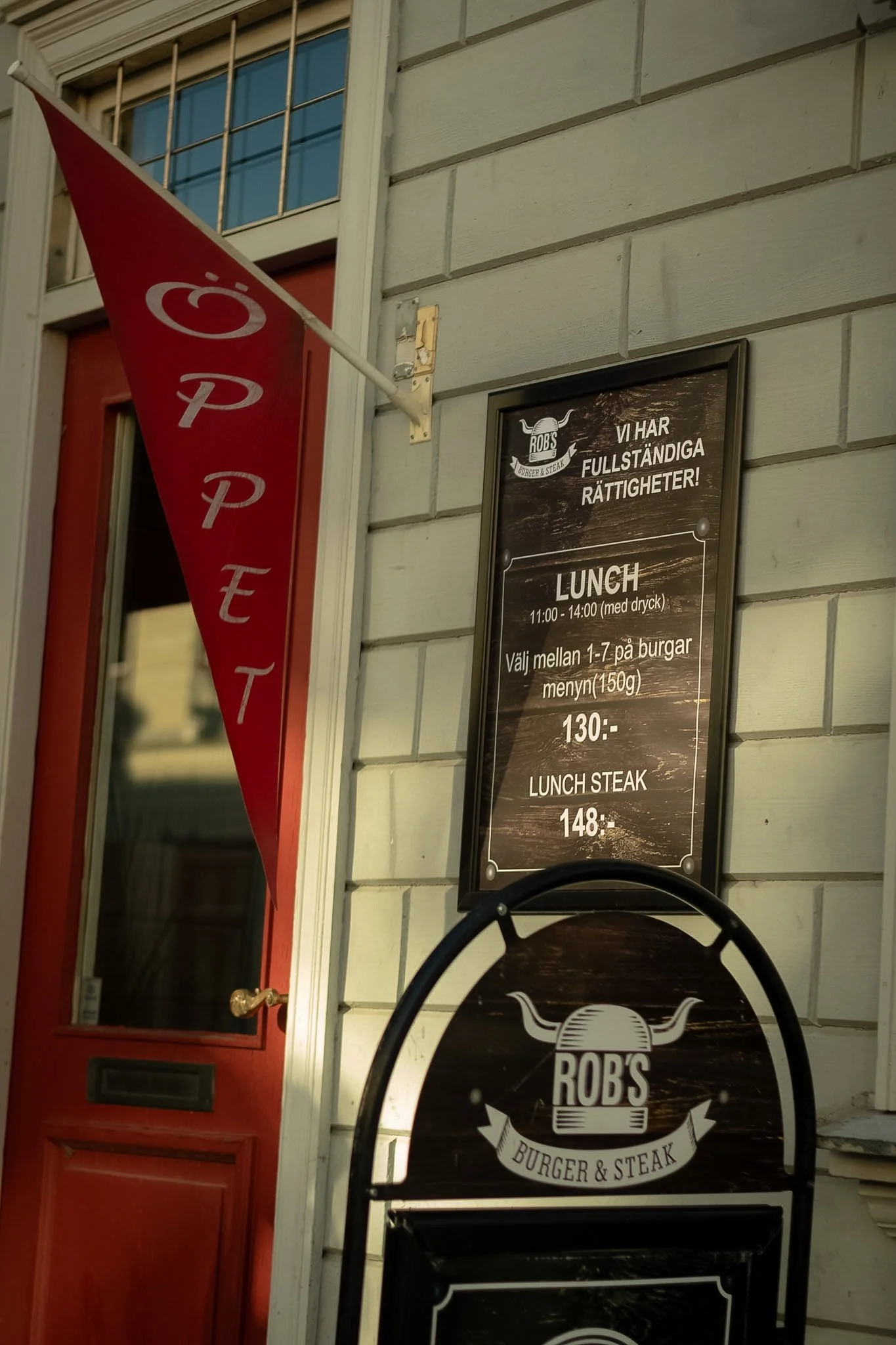 Exterior of a restaurant or burger place with a red door, a window above it, a red vertical sign with white text, and black chalkboard menu signs on the wall. The signs advertise lunch options, including burger and steak meals.