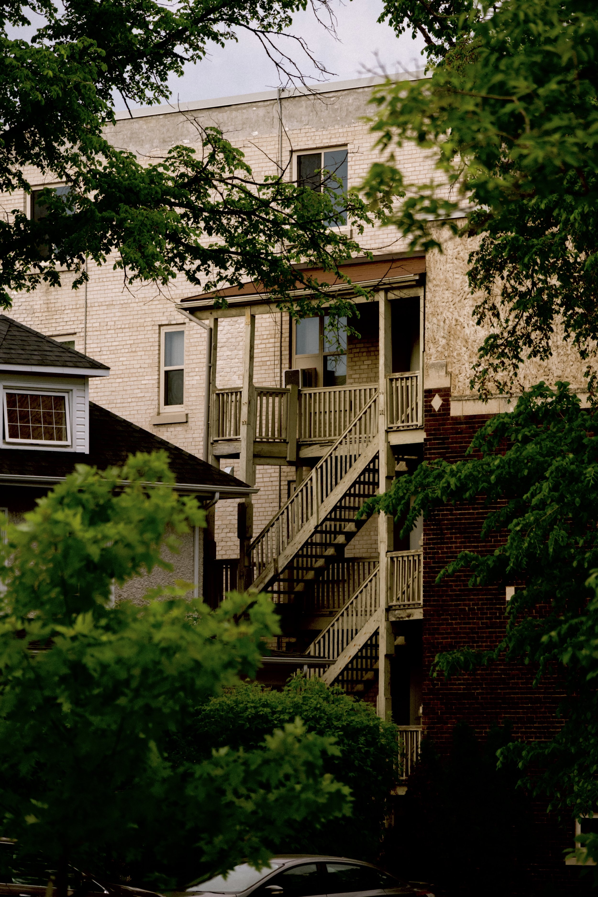 A multi-story residential building with a wooden staircase and a small balcony, partially obscured by green trees and foliage, with a car visible at the bottom of the image.