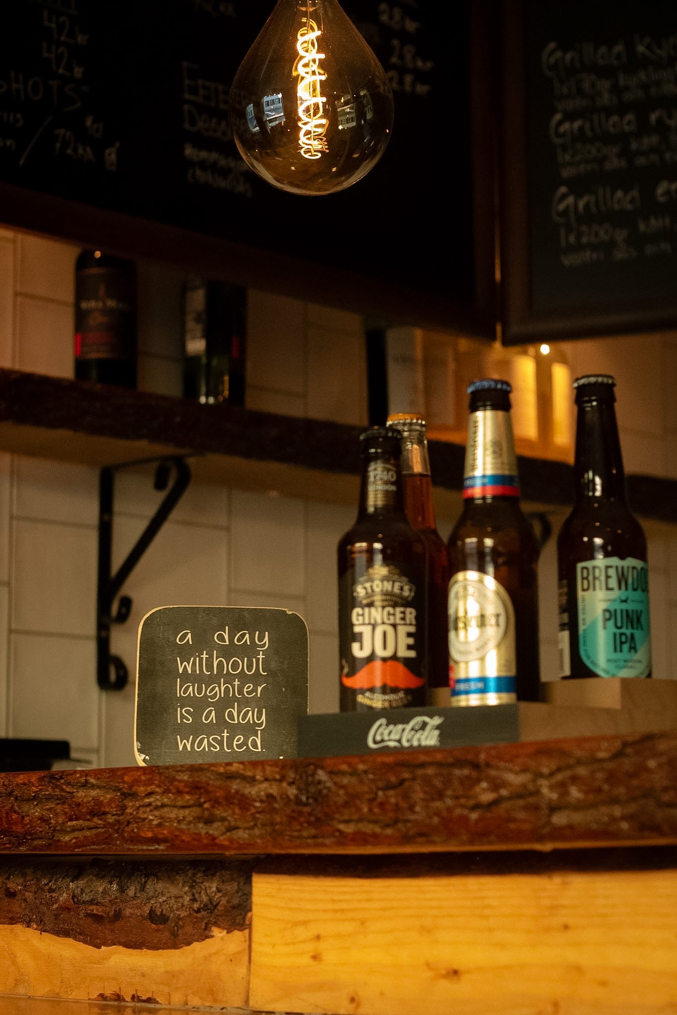 A hanging decorative light bulb with a spiral filament in a dimly lit space above a shelf with bottled beverages, including ginger beer, in a bar or restaurant setting. There is a chalkboard menu and a small sign with an inspirational quote.