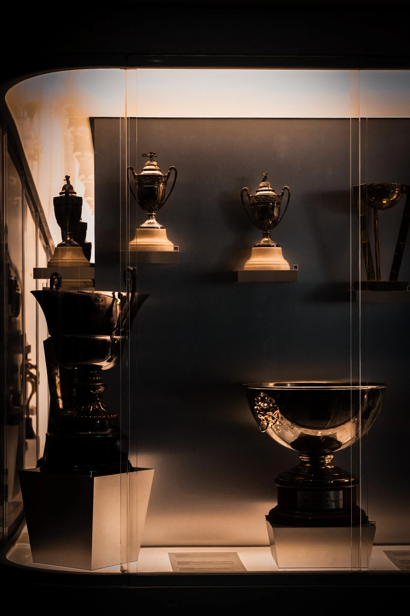 Display case with four vintage silver trophies, two on the top shelf and two on the bottom, in a museum or exhibition setting.