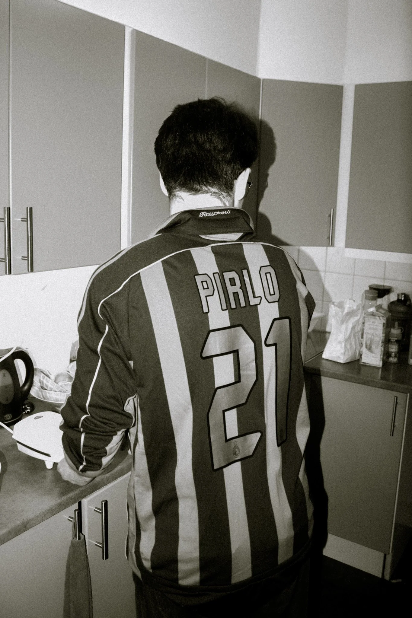 A person with short dark hair wearing a black and white striped soccer jersey with the name PITLO and number 21 on the back, standing at a kitchen counter with cabinets above. The person is facing away from the camera, working or preparing something 