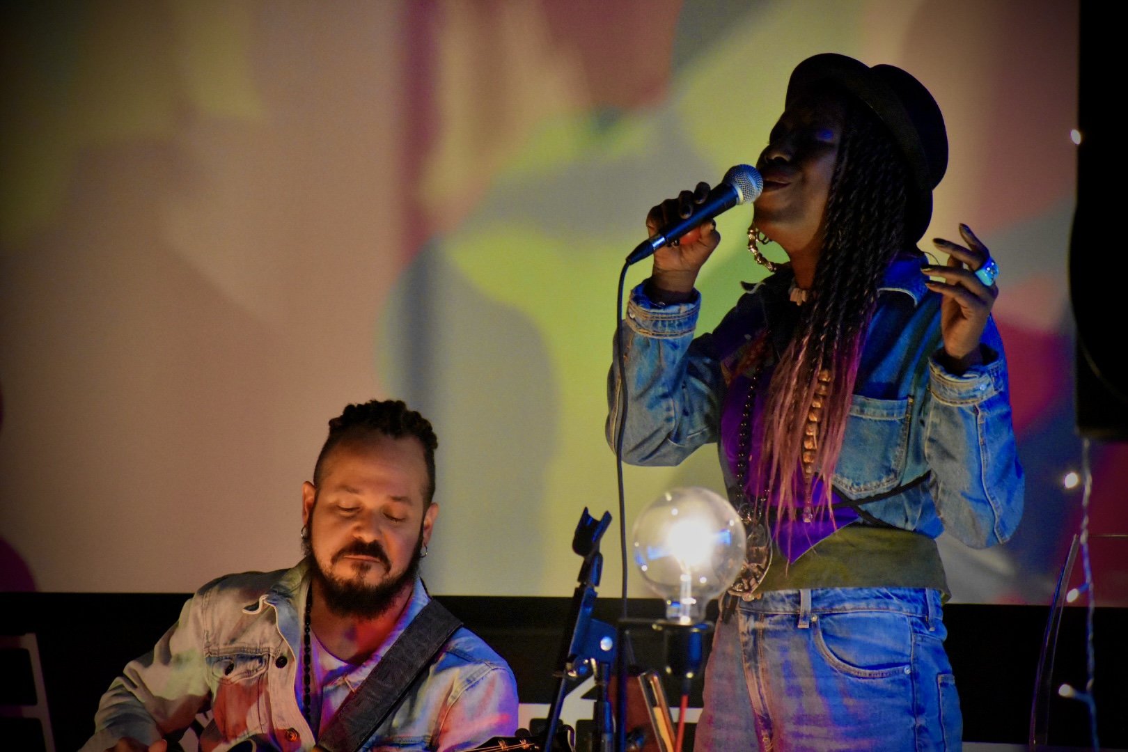 A woman singing into a microphone while wearing a black hat and denim jacket, standing near a man playing an instrument, with colorful blurred background and glowing light in the foreground.