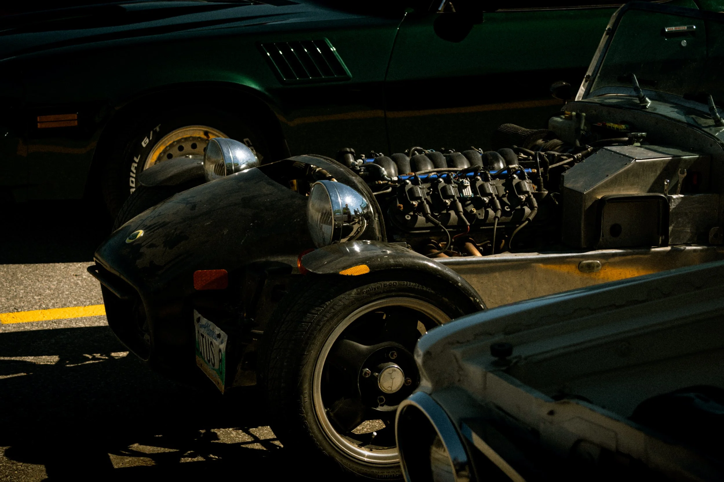 Close-up of a racing car with exposed engine components parked in a lot with other vehicles.
