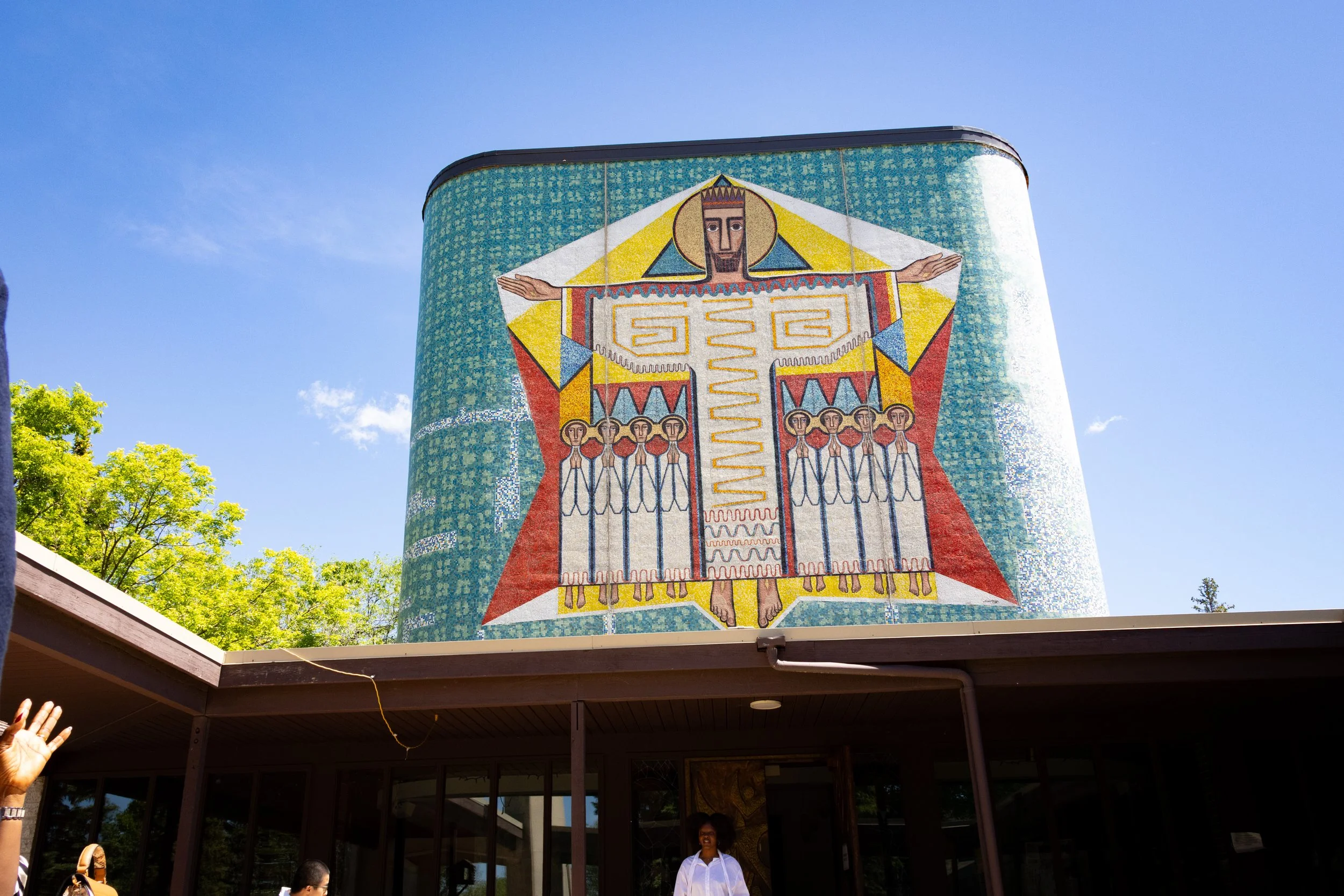 Colorful mosaic mural depicting a figure with outstretched arms, a crown, and multiple smaller figures below, on a tall building against a blue sky.