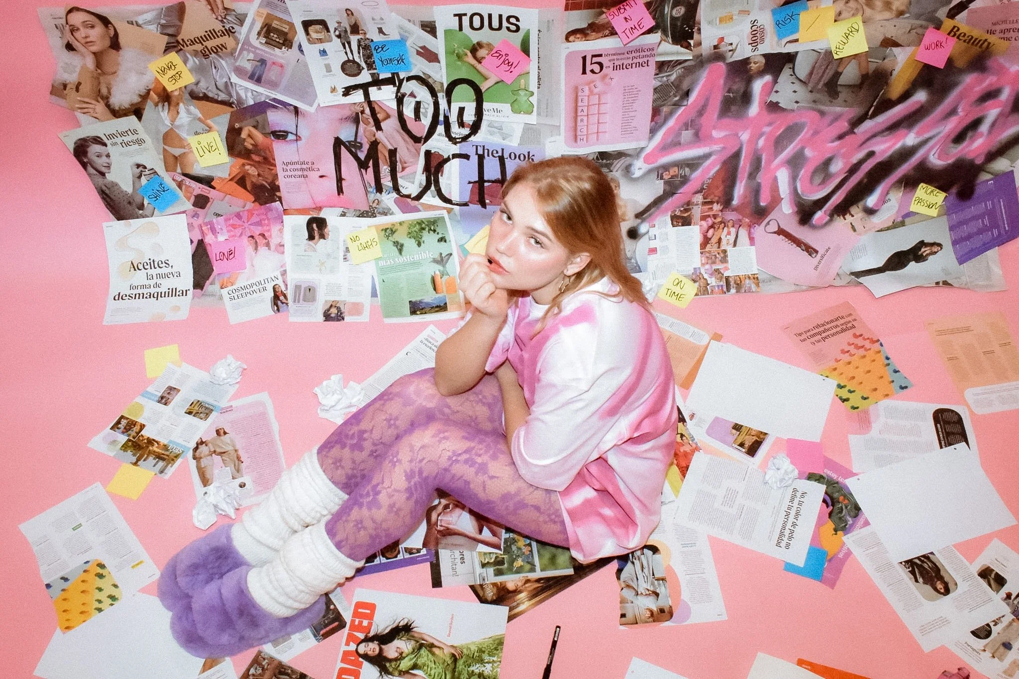 A woman with reddish hair and fair skin, wearing a pink and white tie-dye shirt, purple pants, and white knee-high socks, sits on the floor surrounded by scattered magazines, crumpled tissues, and colorful sticky notes on a pink background.