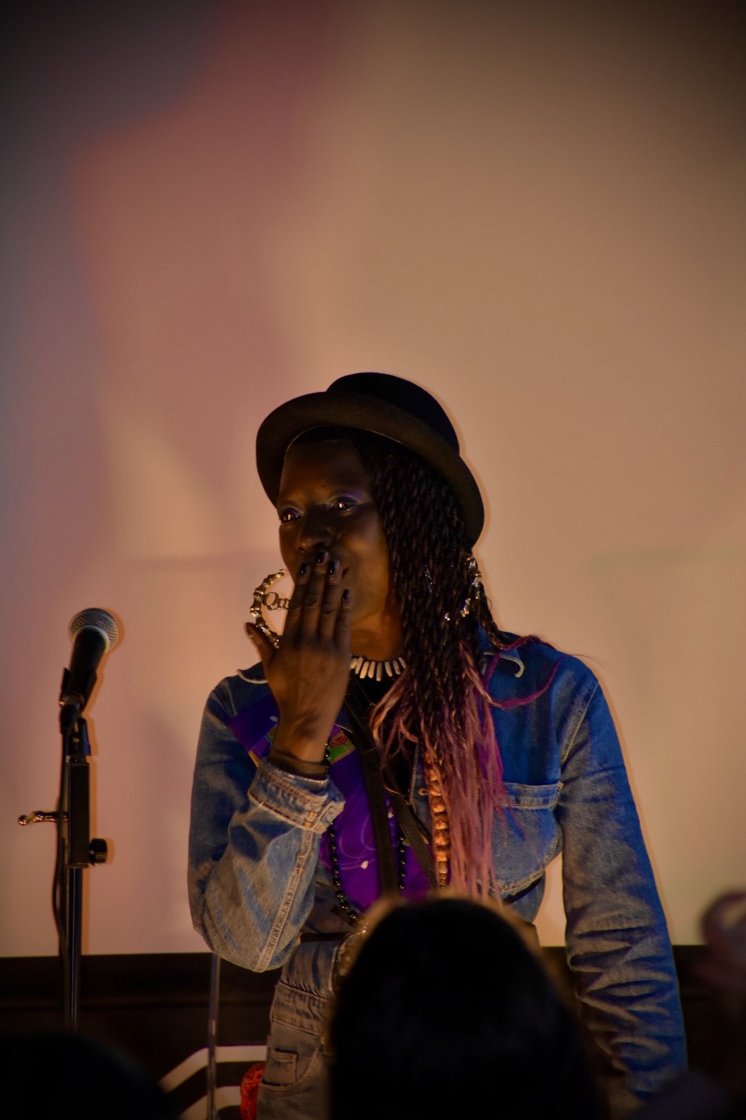A woman with long braided hair wearing a black hat, denim jacket, and colorful accessories standing in front of a microphone, covering her mouth with her hand.
