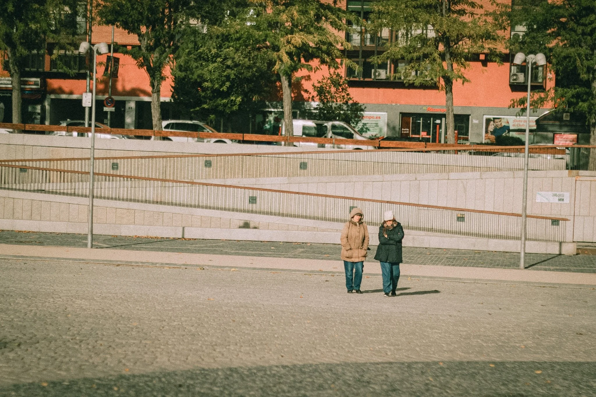 Two women standing and talking in an urban outdoor area with a stone wall, metal railing, and a sidewalk. Behind them, there are parked cars, trees, and a multi-story red brick building with shop fronts.