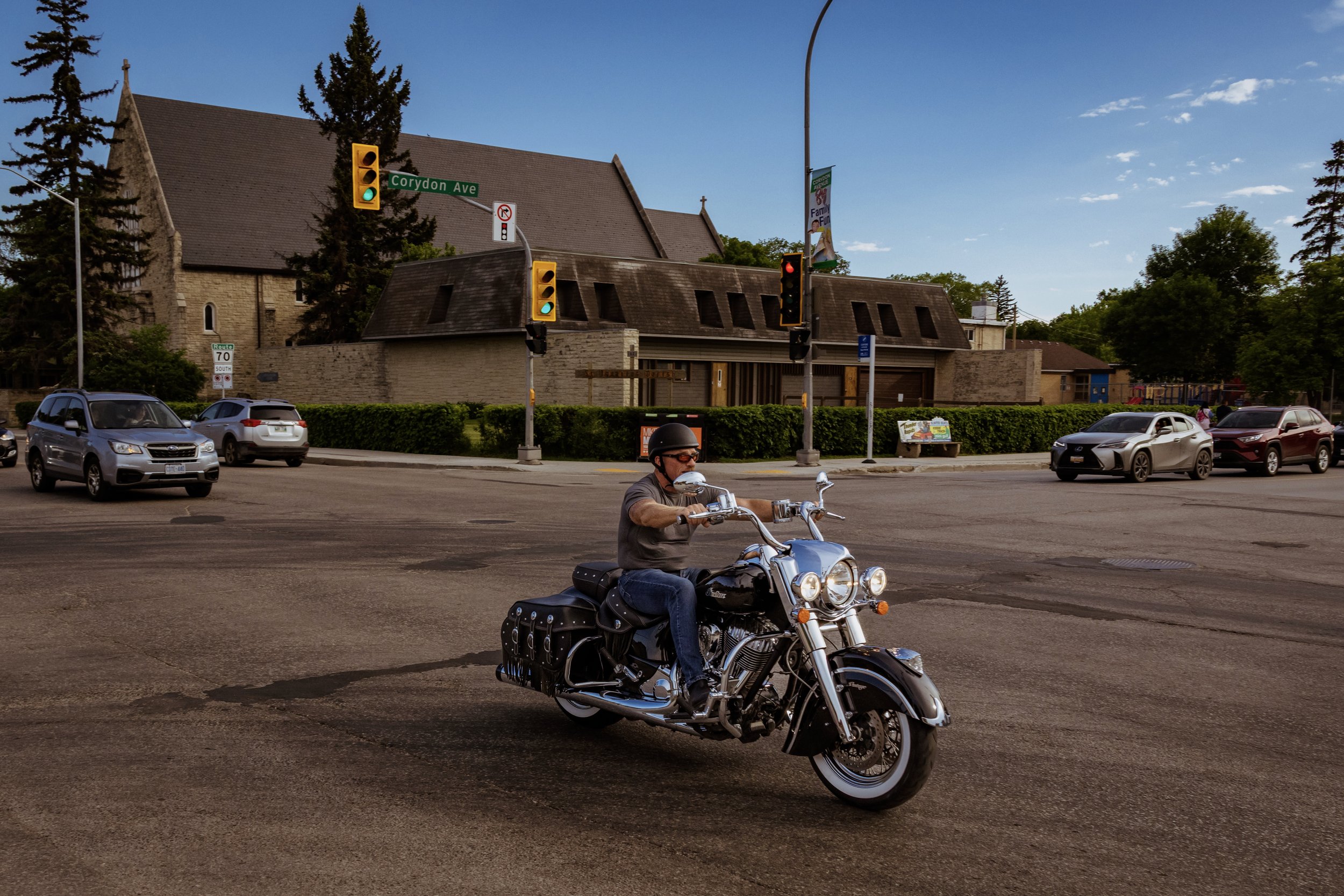 A man riding a black motorcycle with chrome details at an intersection, wearing a black helmet and sunglasses, with multiple cars stopped at the traffic light and a church in the background.