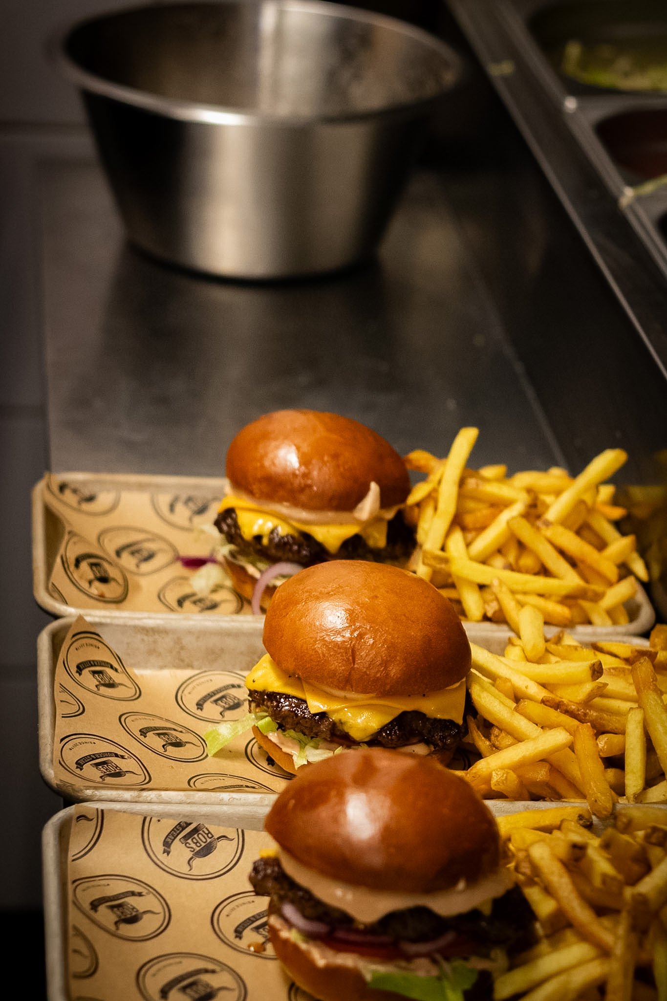 Three cheeseburgers with buns, cheese, lettuce, onions, and beef patties on branded paper trays, with a side of French fries in the background, on a metal surface.