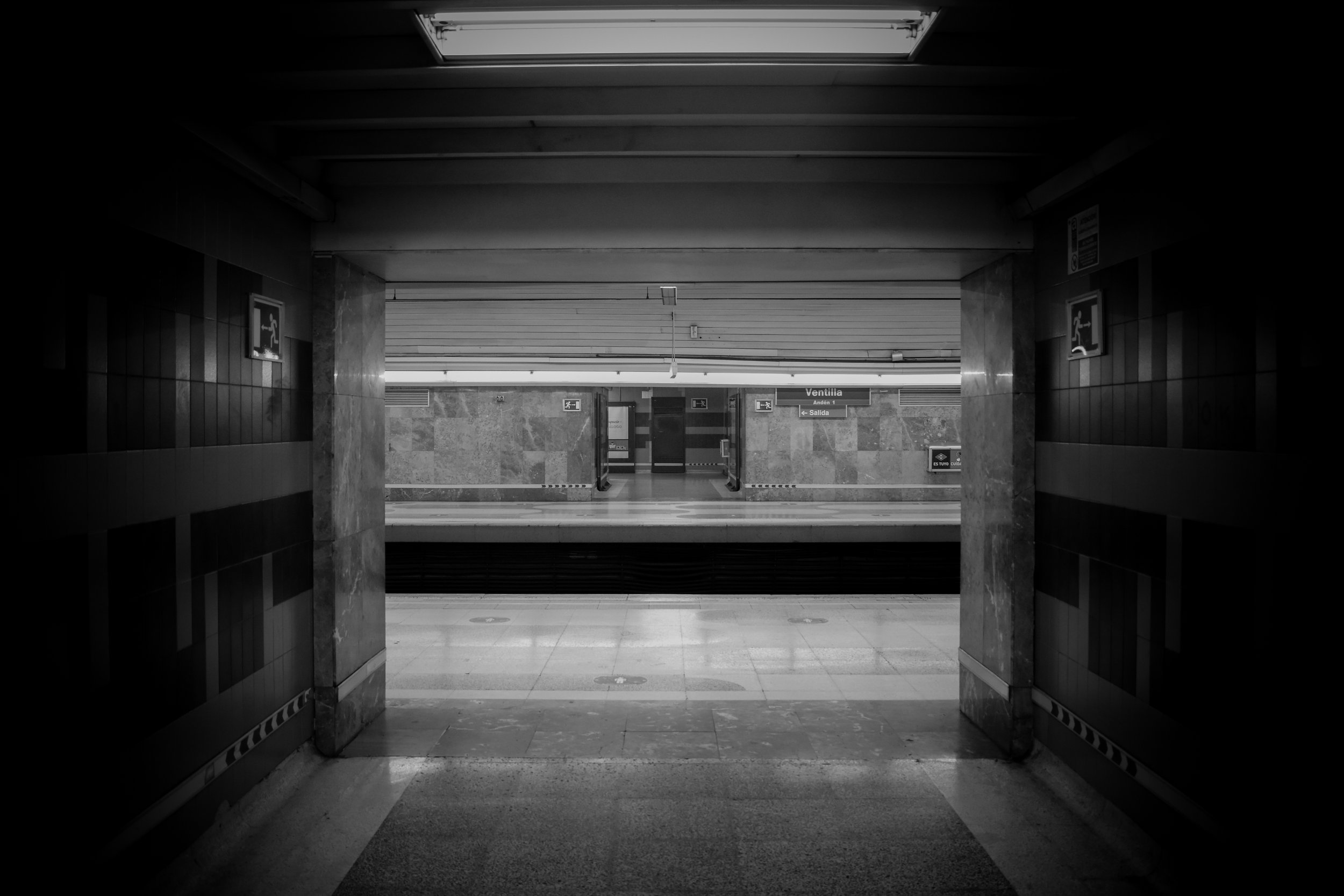 View of an underground metro station entrance with an open passage leading to a platform area, with signs indicating directions to Ventilla and exit, and an emergency exit sign on the left wall.